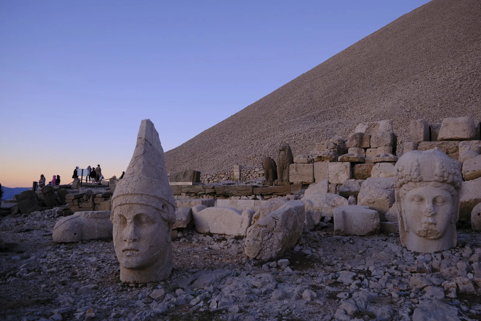 Two stone heads stand in the foreground of a summit terrace, with visitors, animal statues, and a gravel mound behind them.