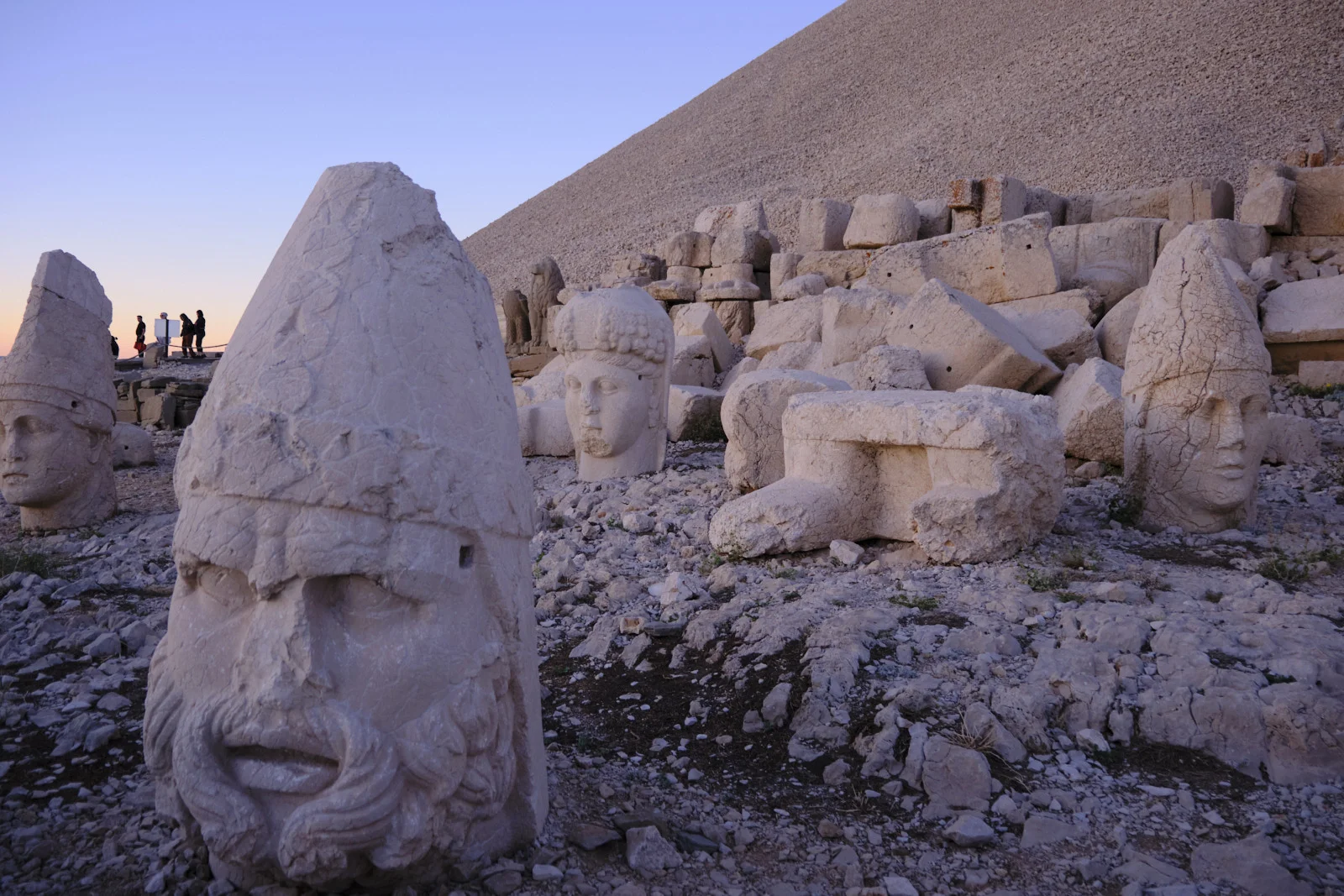 A close bearded stone head in a tall cap stands among other colossal heads and rubble at the base of a gravel mound.