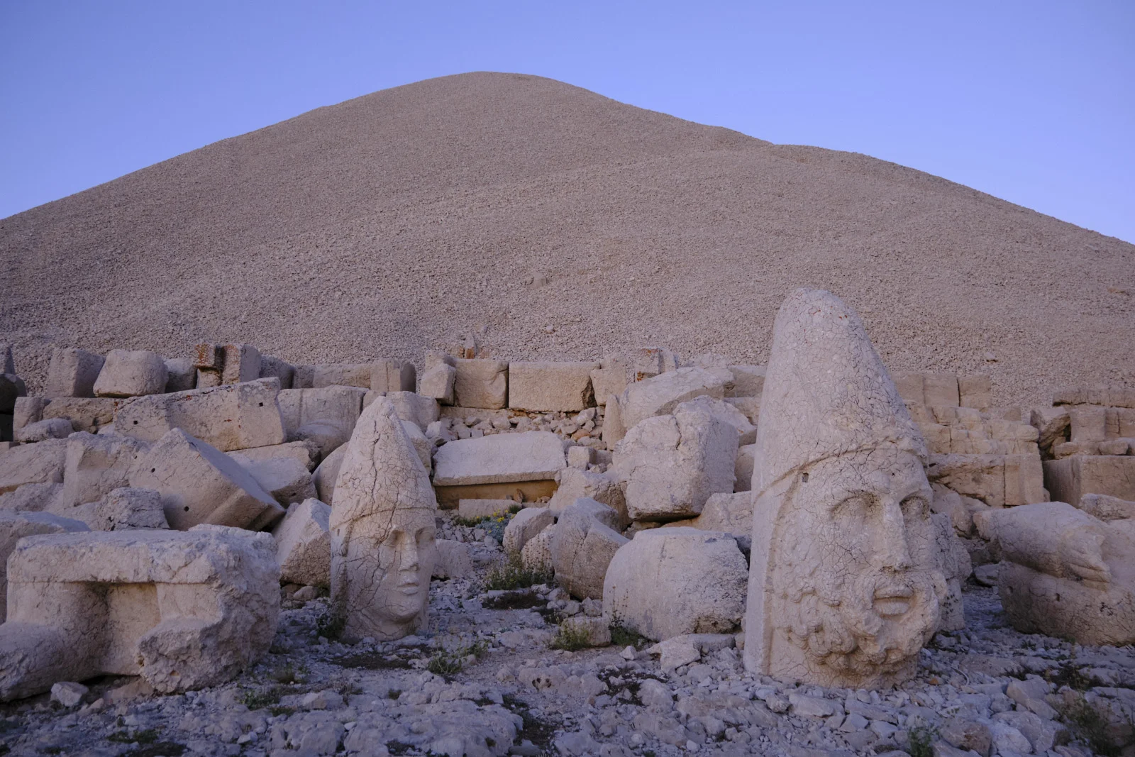 Two colossal stone heads, one bearded with a tall headdress, rest in rubble beneath a huge gravel mound.
