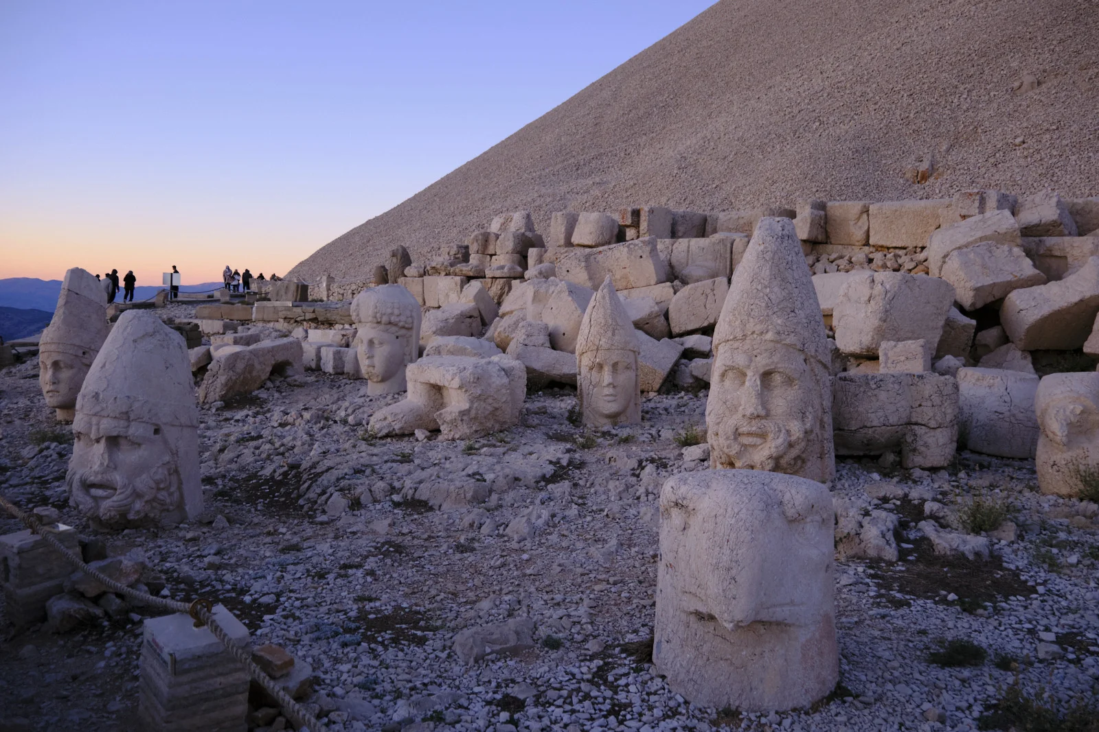 Several colossal stone heads and an eagle stand in rubble beneath a gravel mound, with visitors on the terrace at dusk.