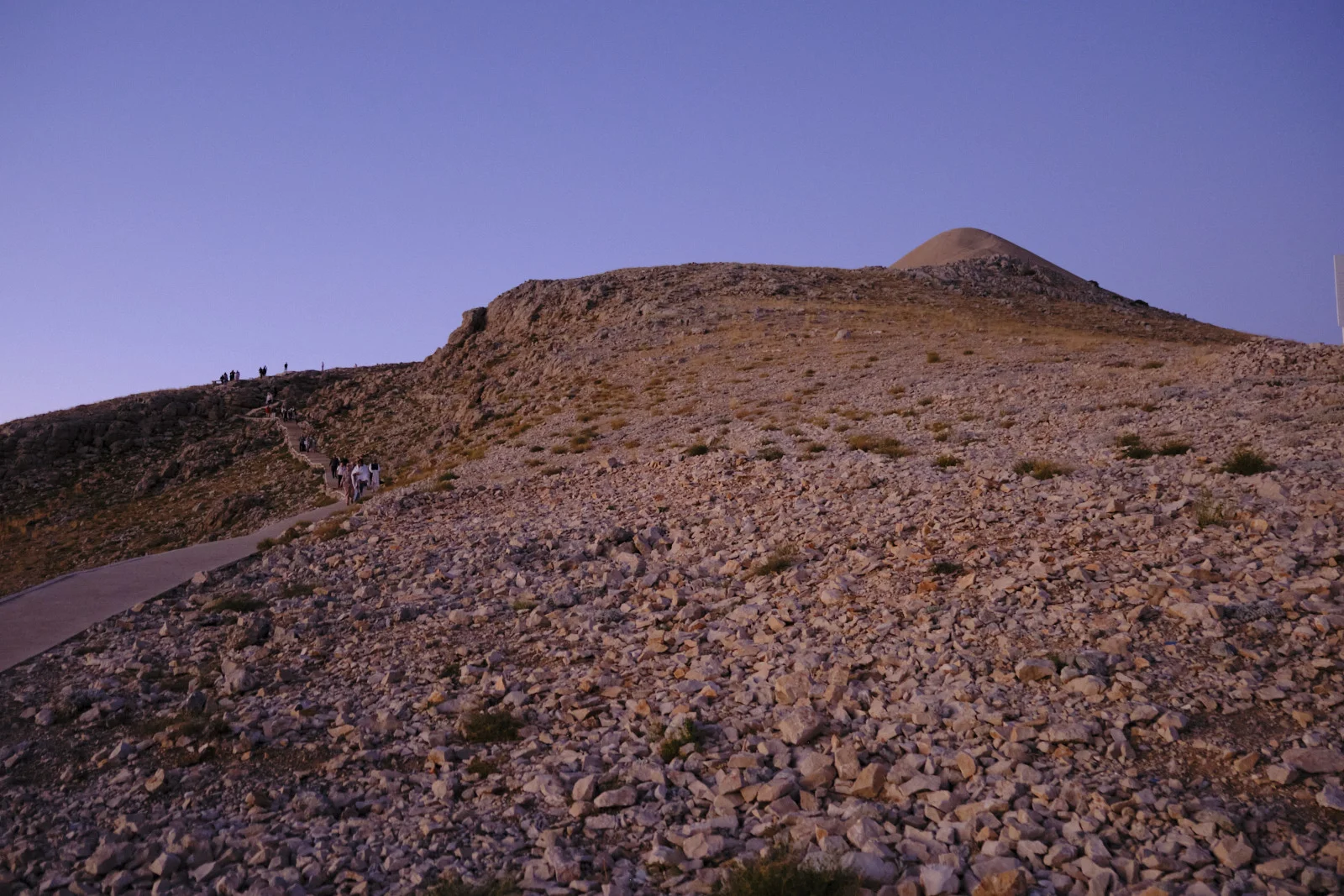 Visitors walk a path down a rocky slope, with the conical gravel tumulus of Mount Nemrut on the ridge behind them.