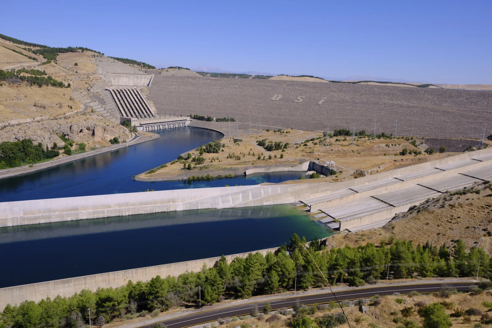 A rockfill dam marked DSİ rises behind a blue canal, a highway, and pine trees under a clear sky.