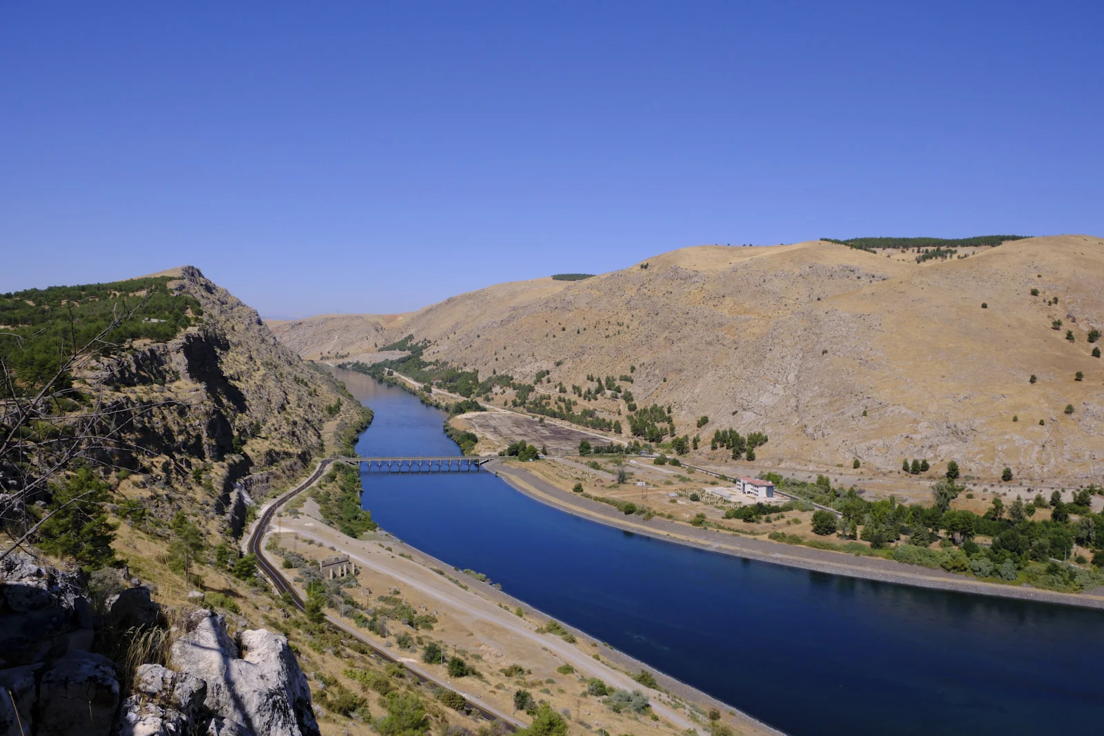 A blue river curves through arid hills, crossed by a low bridge, with a rocky cliff in the foreground.