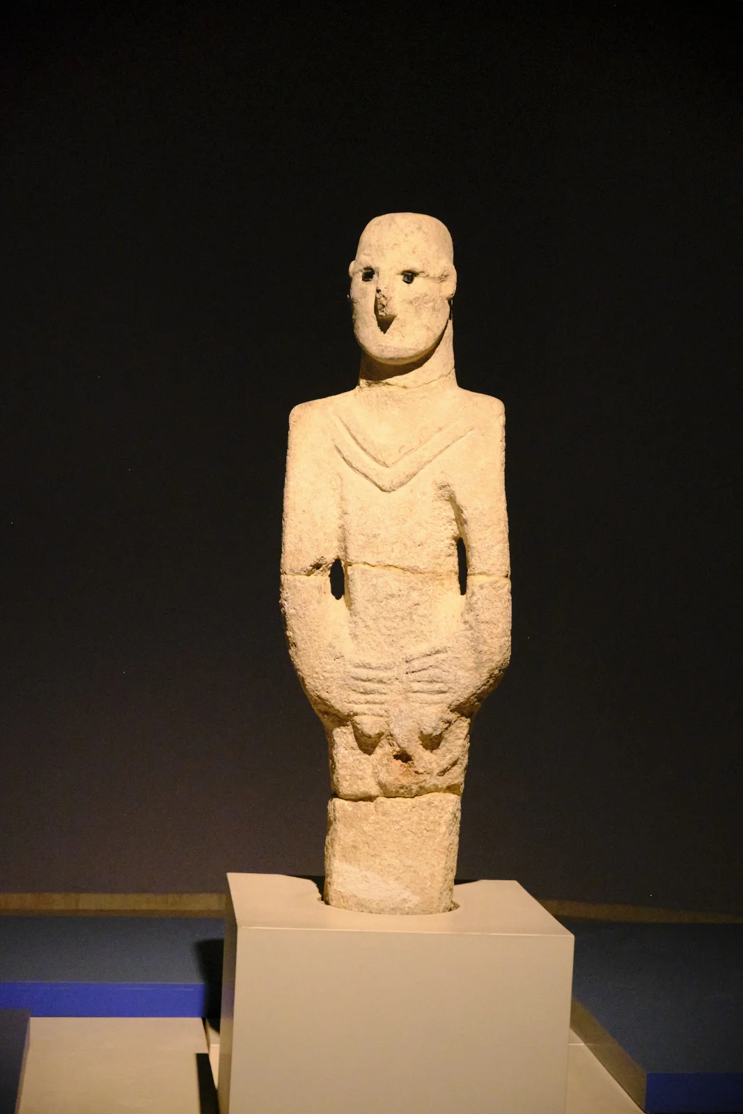 A limestone human statue with dark inlaid eyes and a V-shaped collar stands on a museum pedestal.