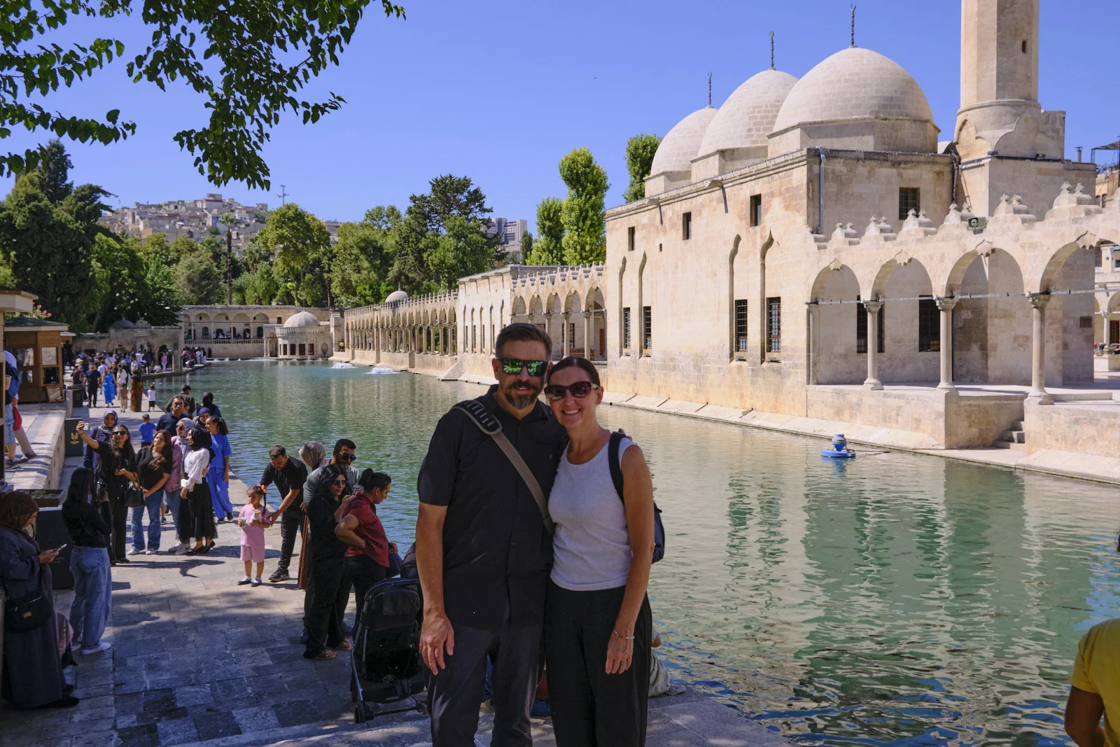 A couple stands by the sacred pool while visitors gather along the walkway and a domed mosque rises behind them.