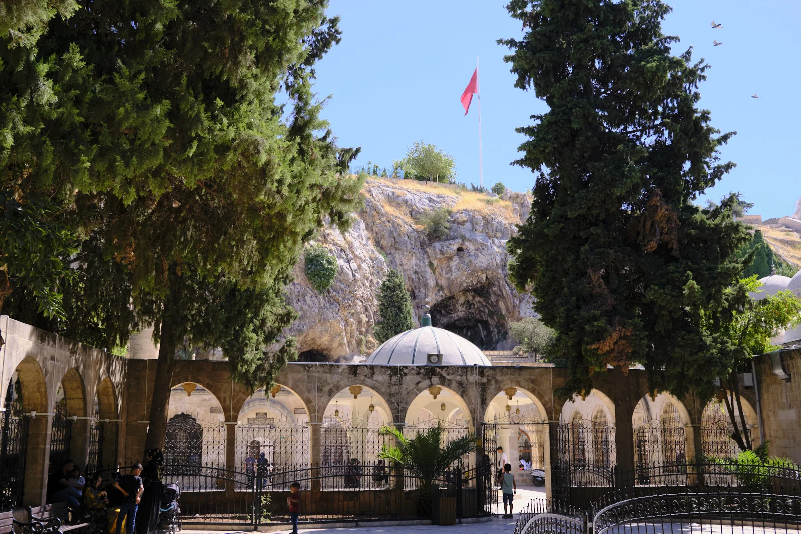 Cave openings in a limestone cliff rise above a white dome, an arched courtyard, and an iron fence, with a Turkish flag on the ridge.