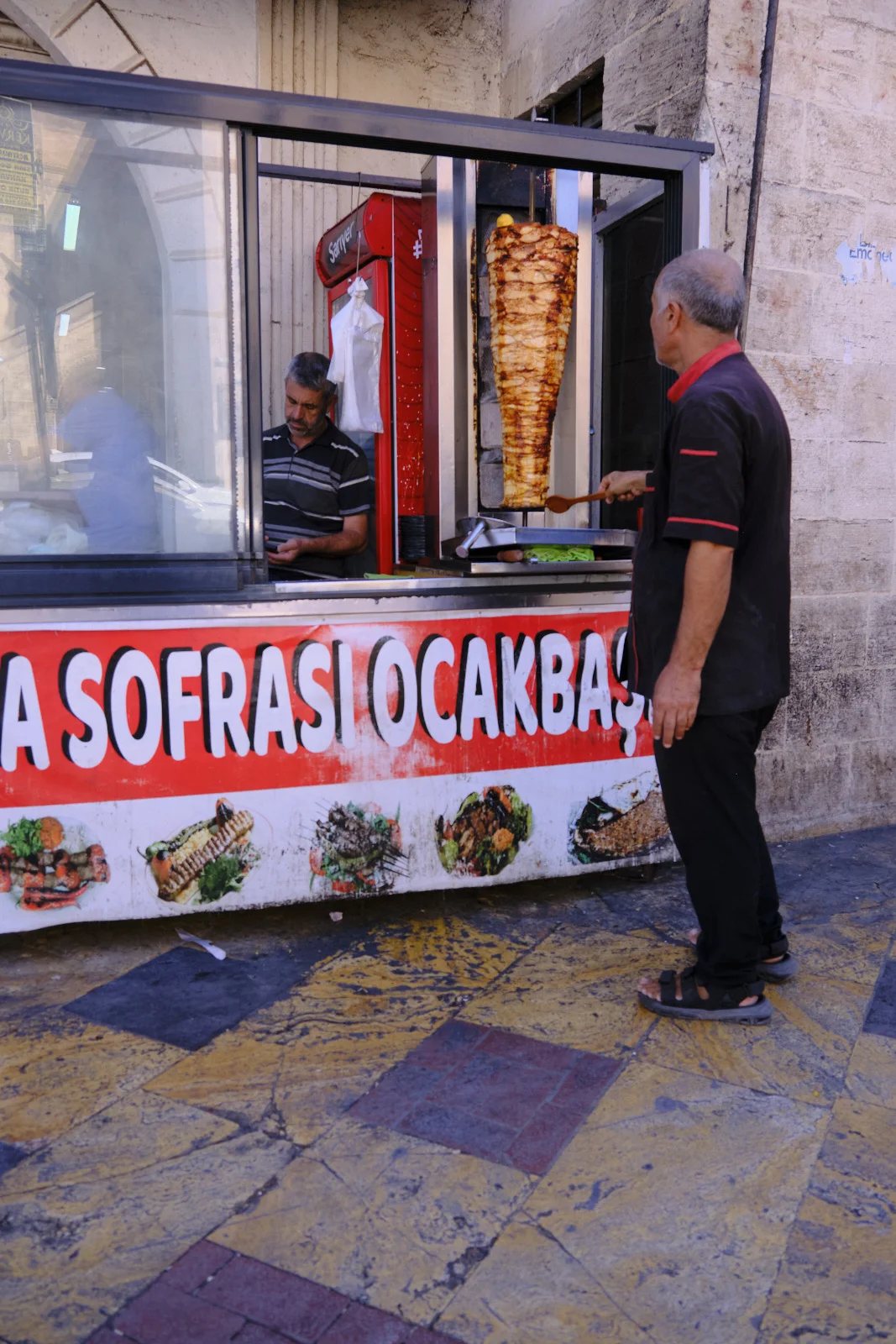 A cook stands beside a tall vertical spit of meat at an outdoor grill stall on a tiled sidewalk.