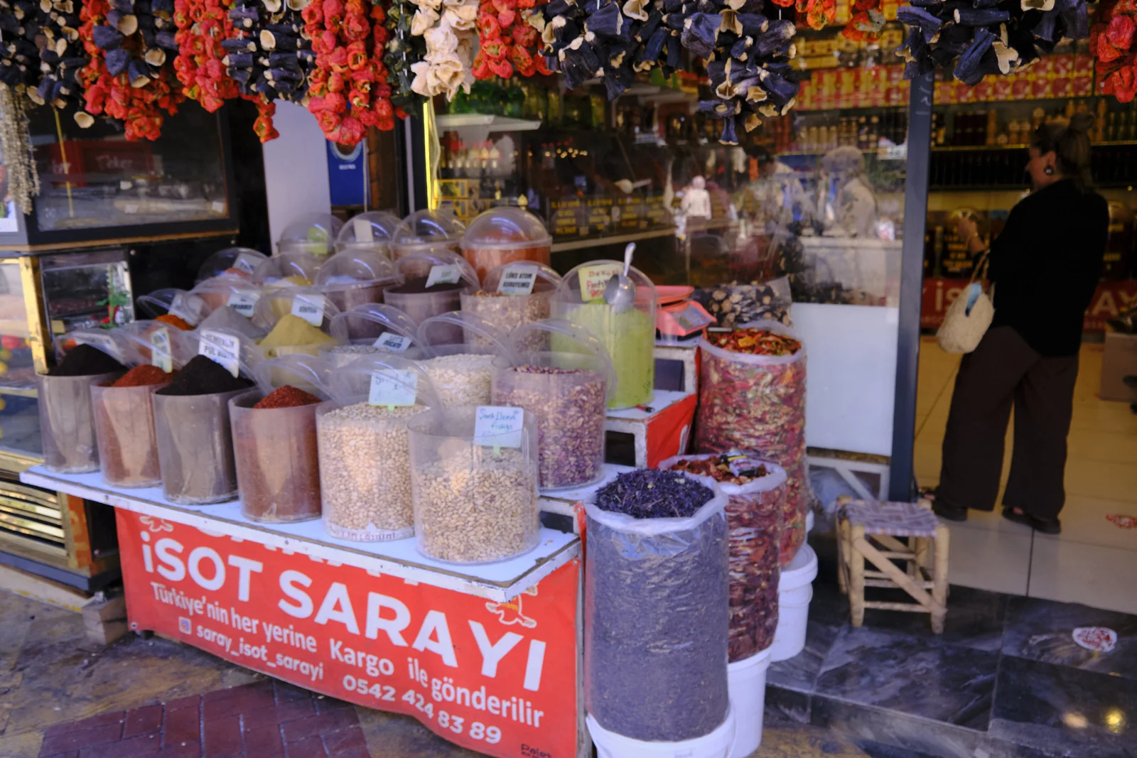 Jars of spices and nuts line a red market counter beneath hanging strings of dried peppers.
