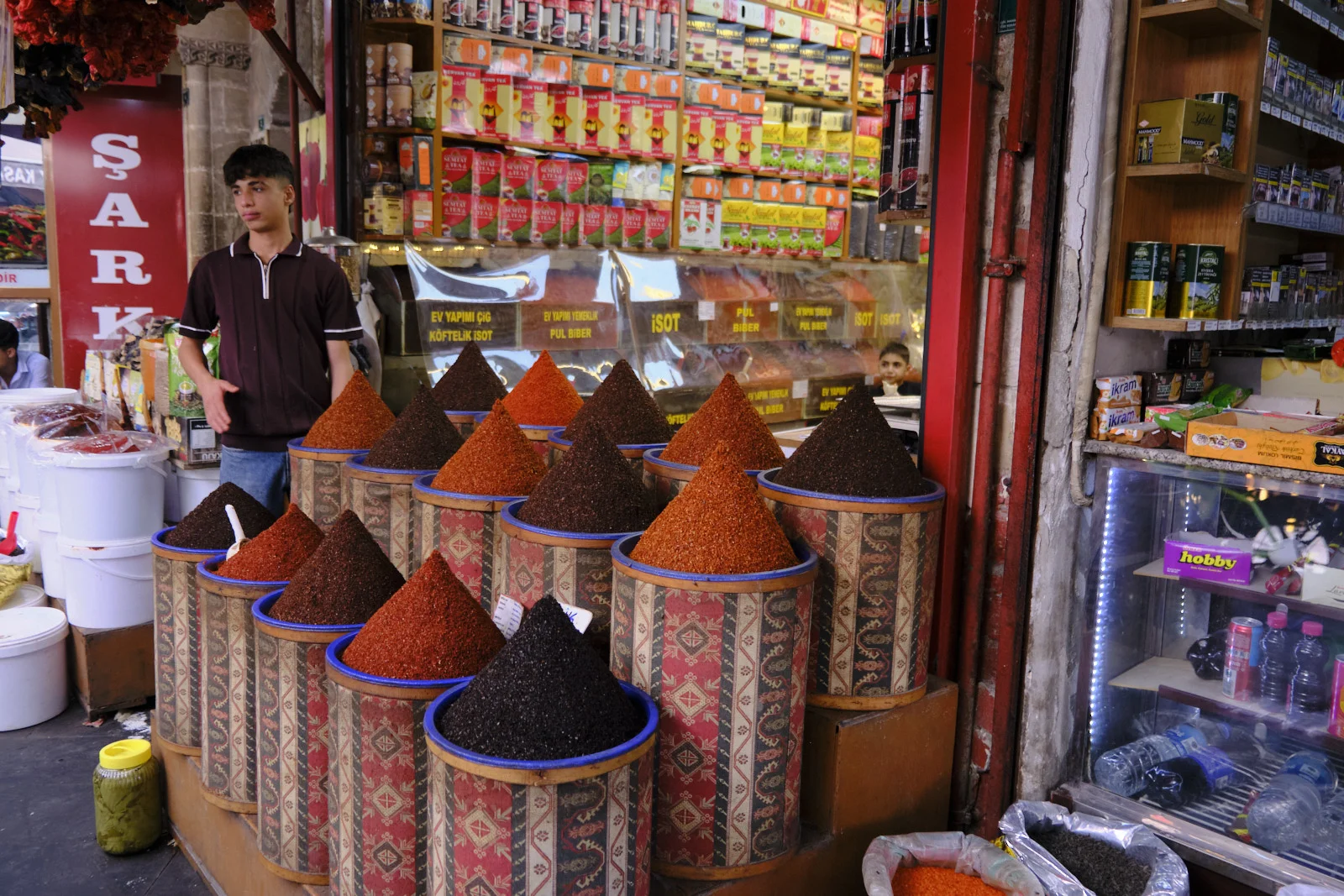 Conical piles of red, orange, and black ground spices fill patterned tins in front of a shopkeeper.