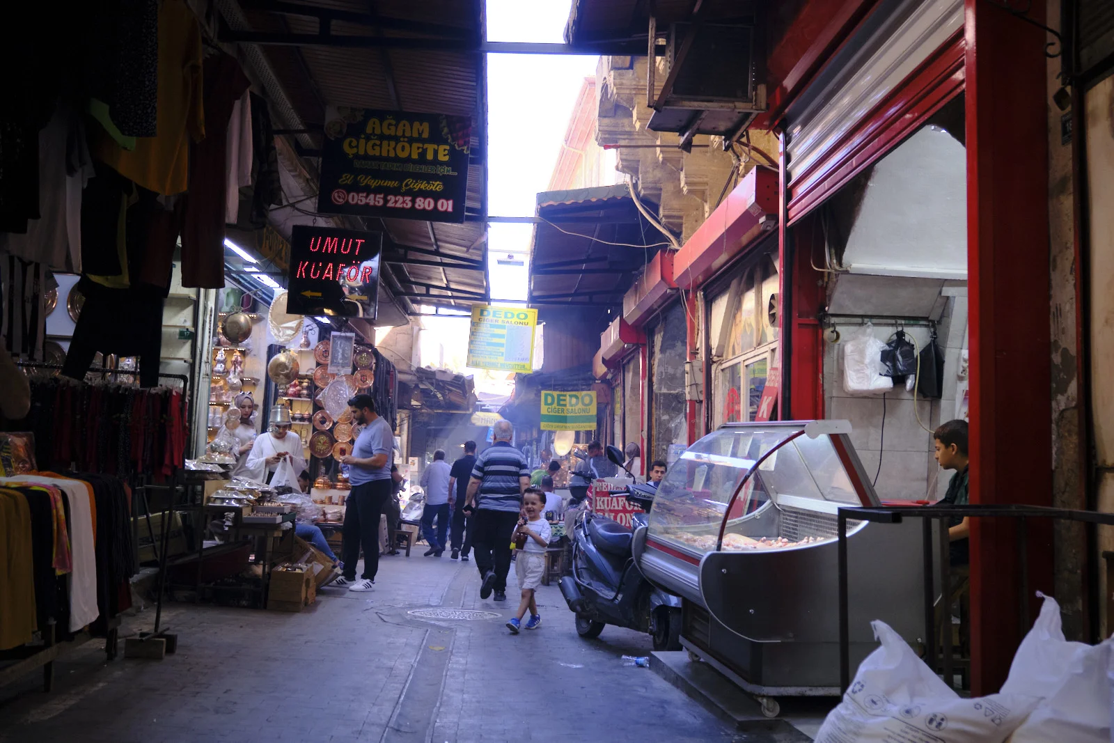 A child walks through bazaar smoke past hanging clothes, copper pans, and a parked scooter.