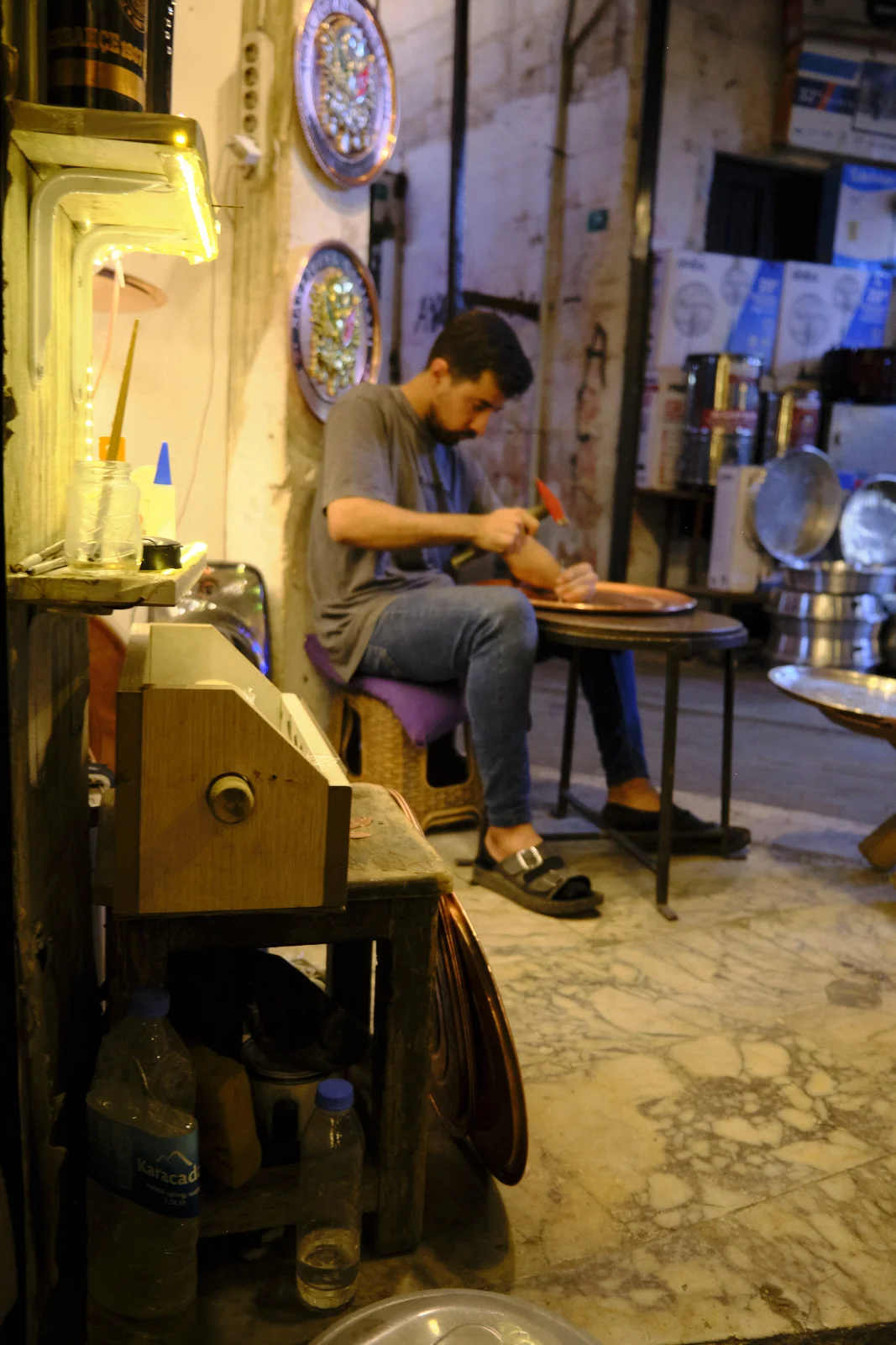 A seated craftsman hammers the rim of a copper tray in a cluttered metal workshop.