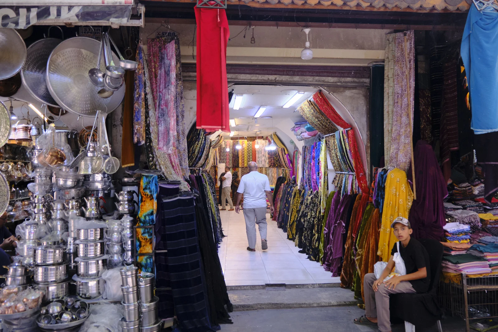 Metal cookware hangs beside a narrow passage lined with colorful fabrics, where a man walks away and a boy sits.
