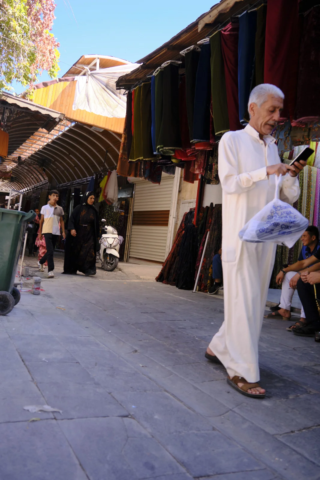 A man in a long white robe checks his phone while walking past hanging garments in a market lane.