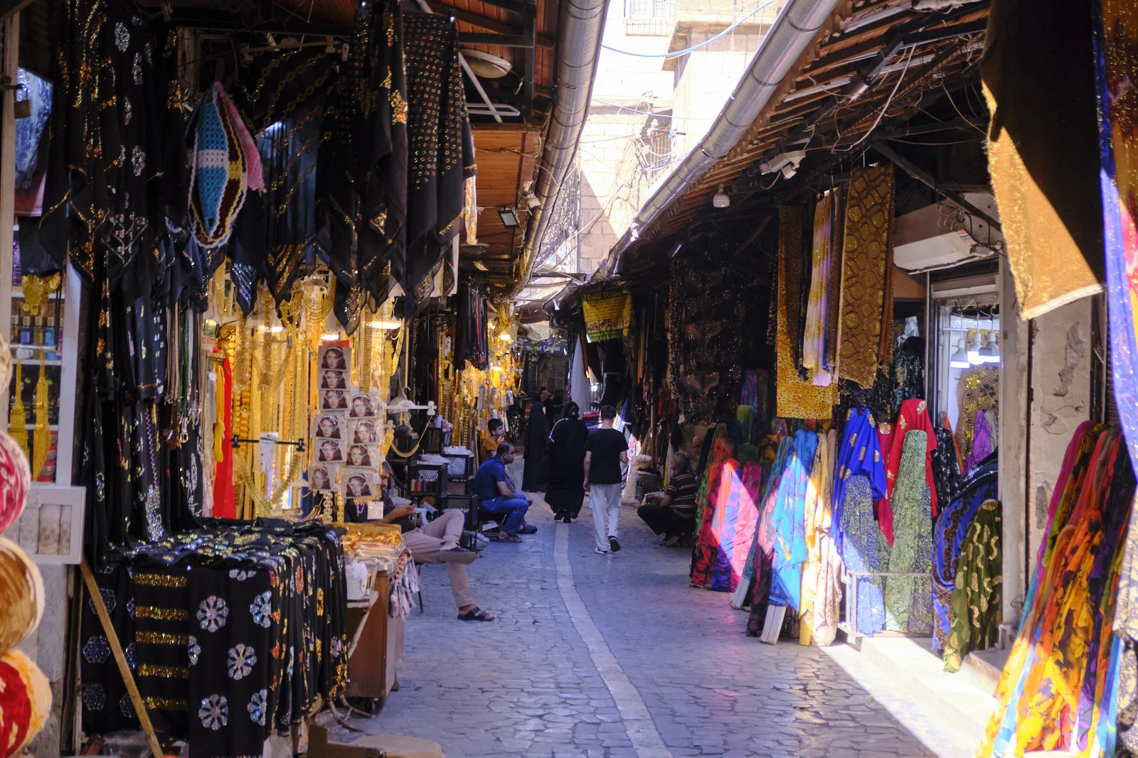 Sequined black garments and brightly colored dresses hang along both sides of a narrow sunlit market lane.