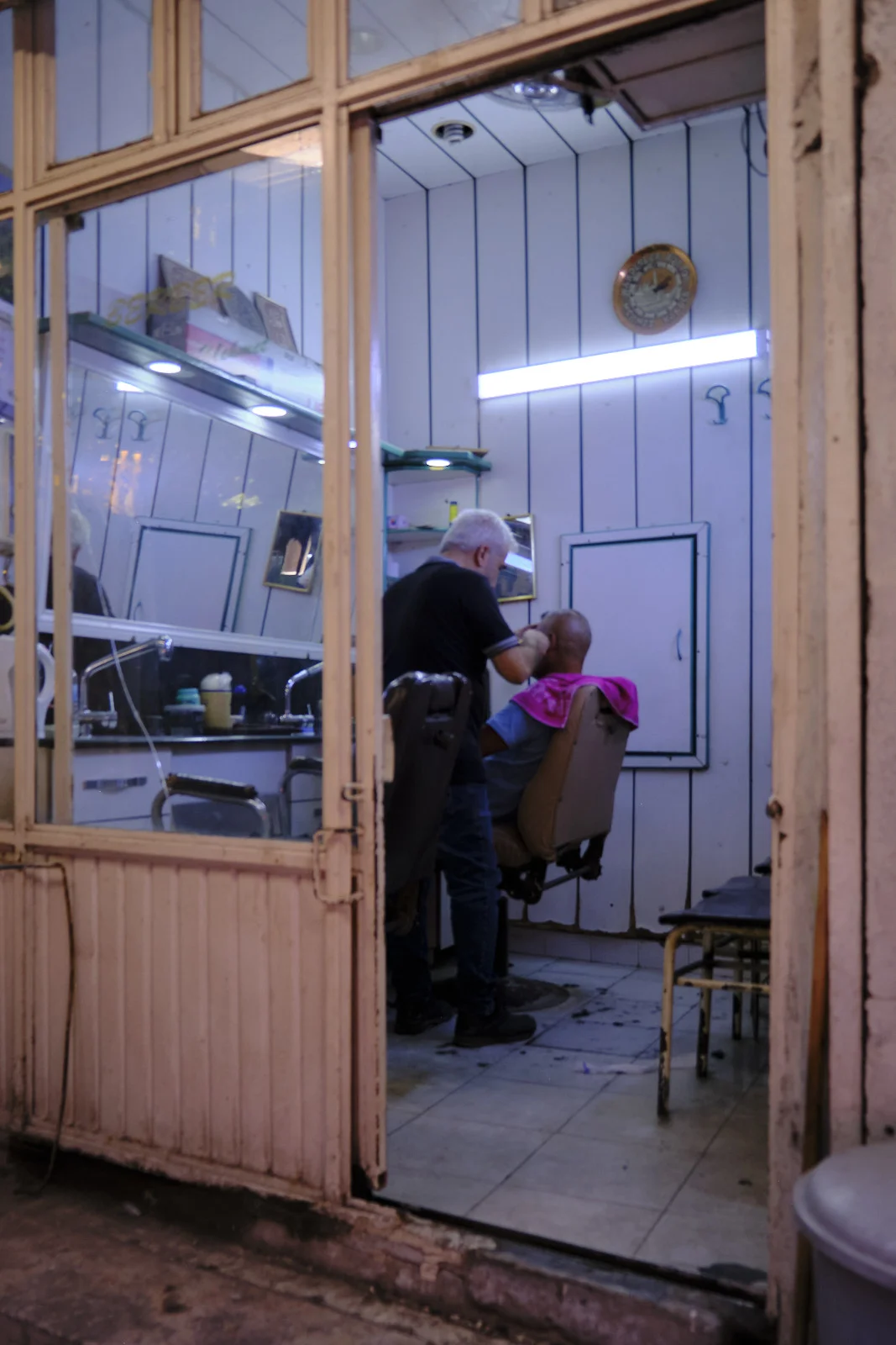 A barber leans over a seated customer inside a small shop seen through an open, worn doorway.