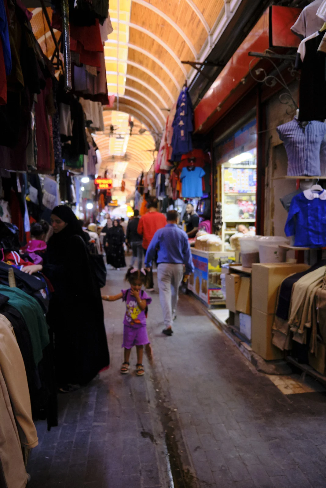 A young girl in purple stands in a crowded covered bazaar beneath repeating wooden arches and hanging clothes.