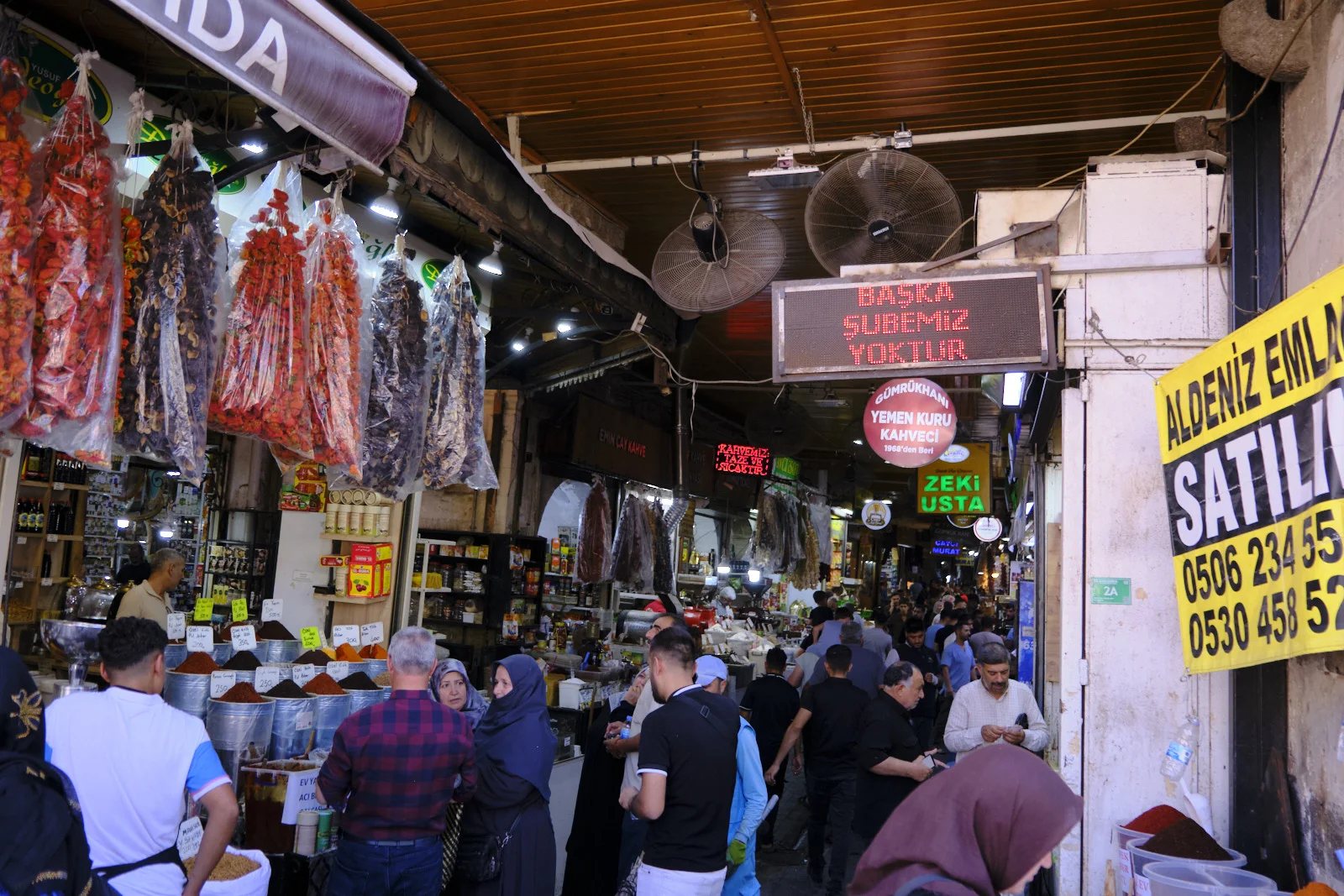 A dense crowd moves through a market hall beneath hanging dried peppers, spice bins, and ceiling fans.