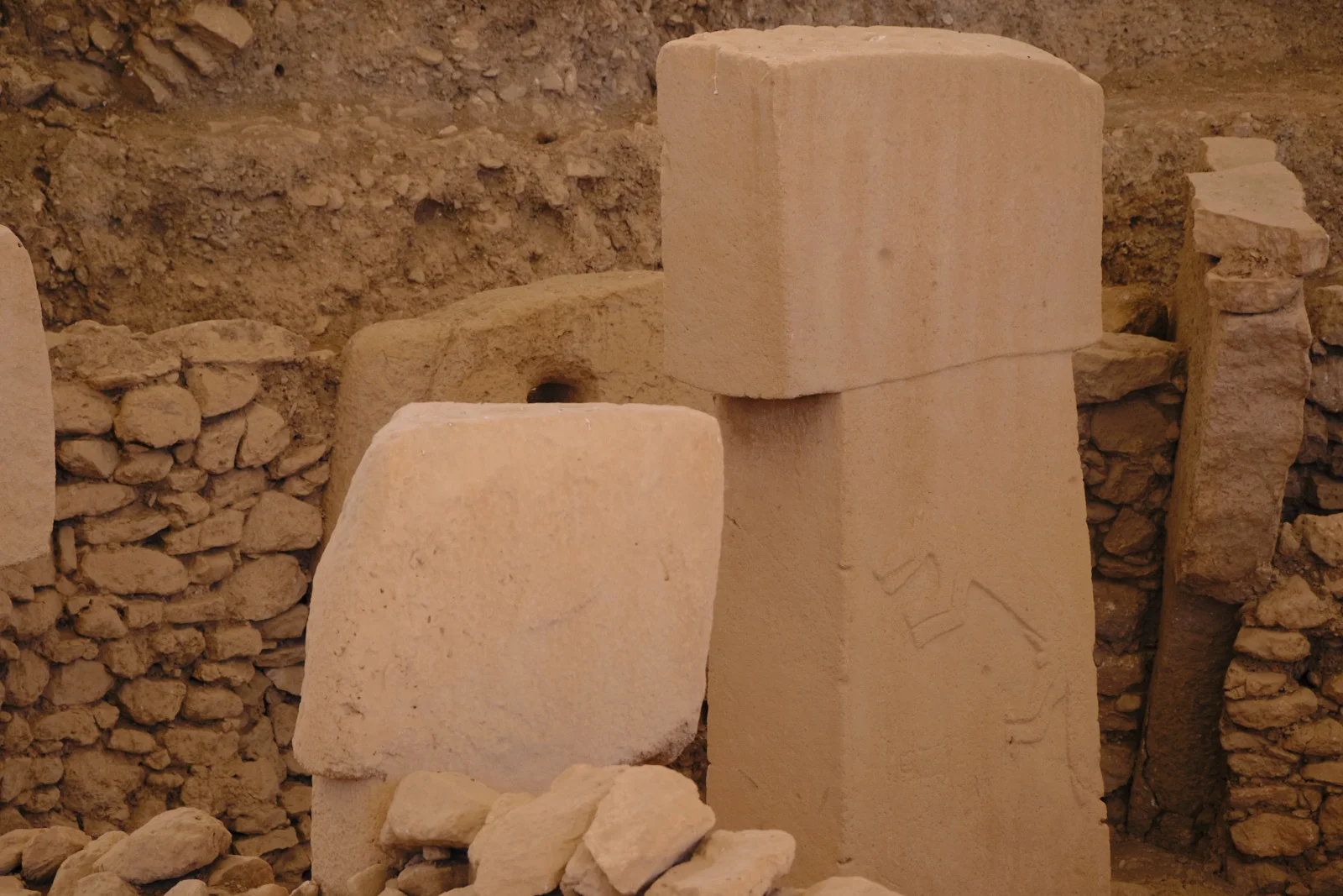 A T-shaped limestone pillar with a faint animal relief stands among rubble walls and a second pillar.