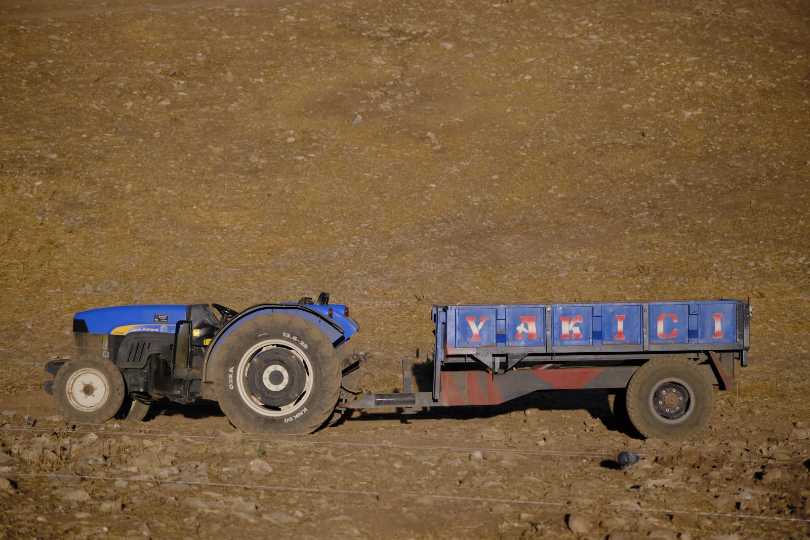 A blue tractor hitched to a blue trailer is parked on a dry, rocky hillside.