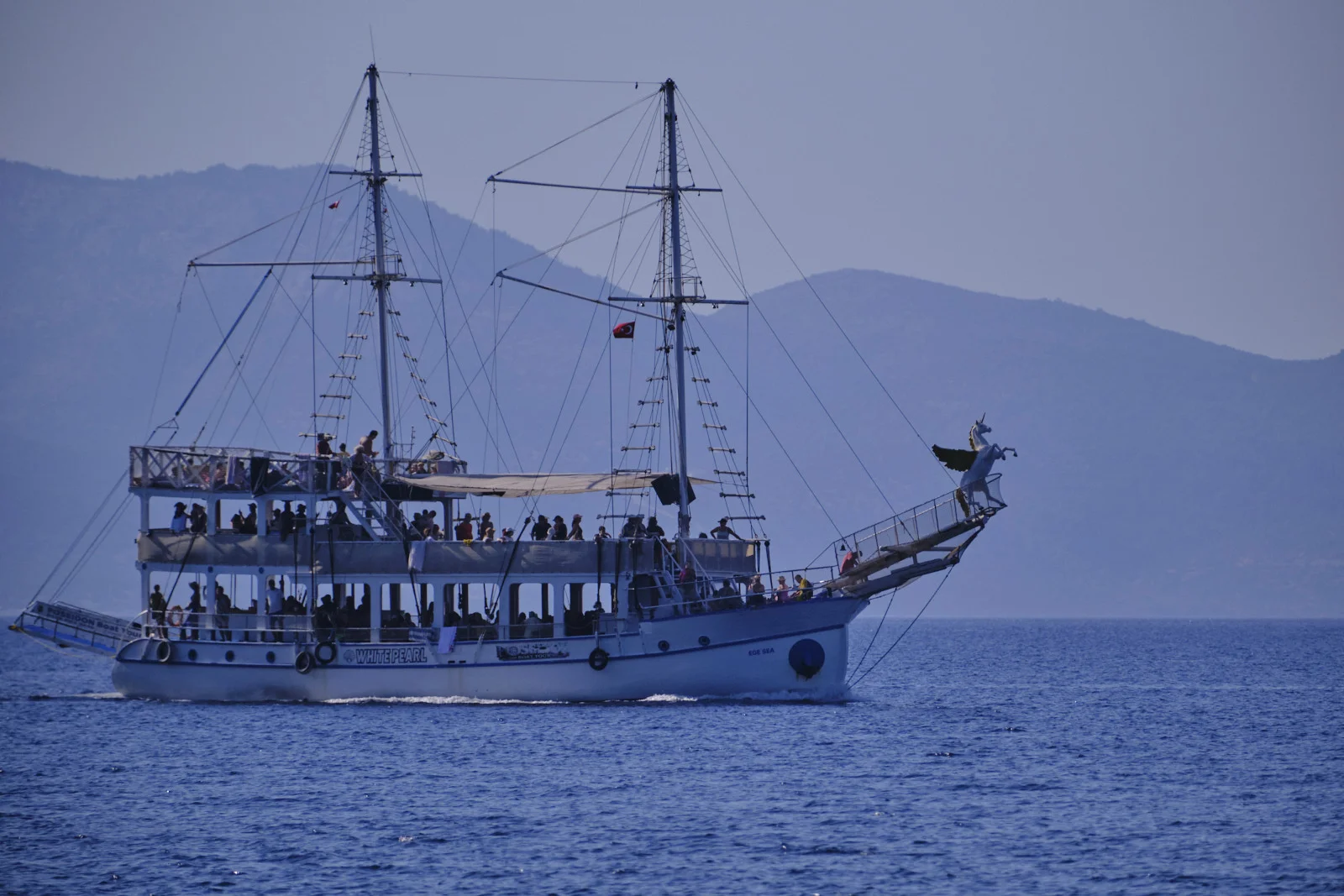 A white two-deck tour boat with tall masts and a winged-horse figurehead sails on deep blue water beneath hazy mountains.