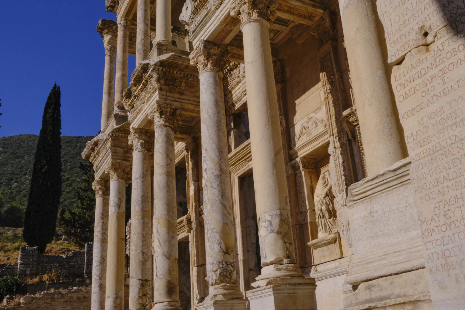 Marble columns and ornate stonework of a classical facade, with a Greek inscription covering the wall at the right, under a deep blue sky.