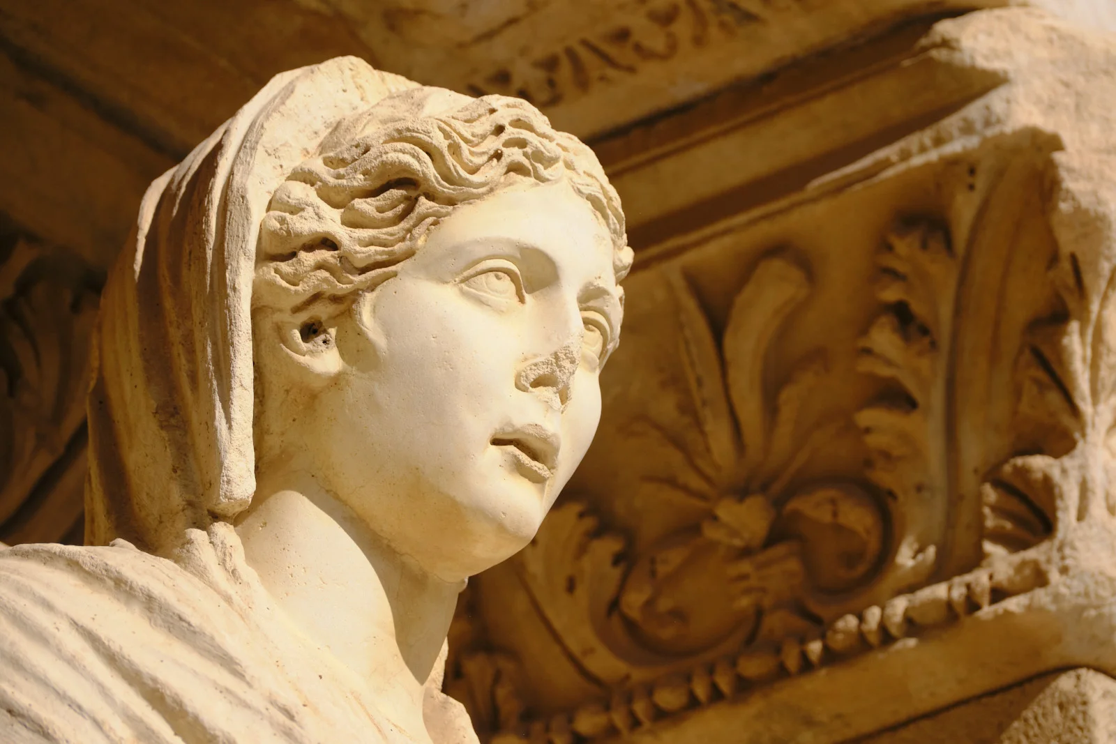 Close view of a weathered marble head with curly hair and a veil, set against a carved Corinthian capital.