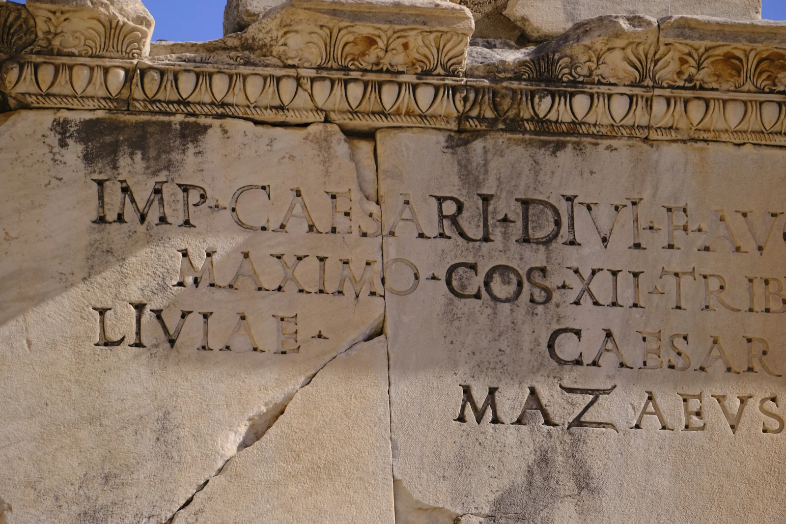 Close view of a cracked marble inscription in Latin letters, including the names Caesar, Livia, and Mazaeus, beneath carved molding.