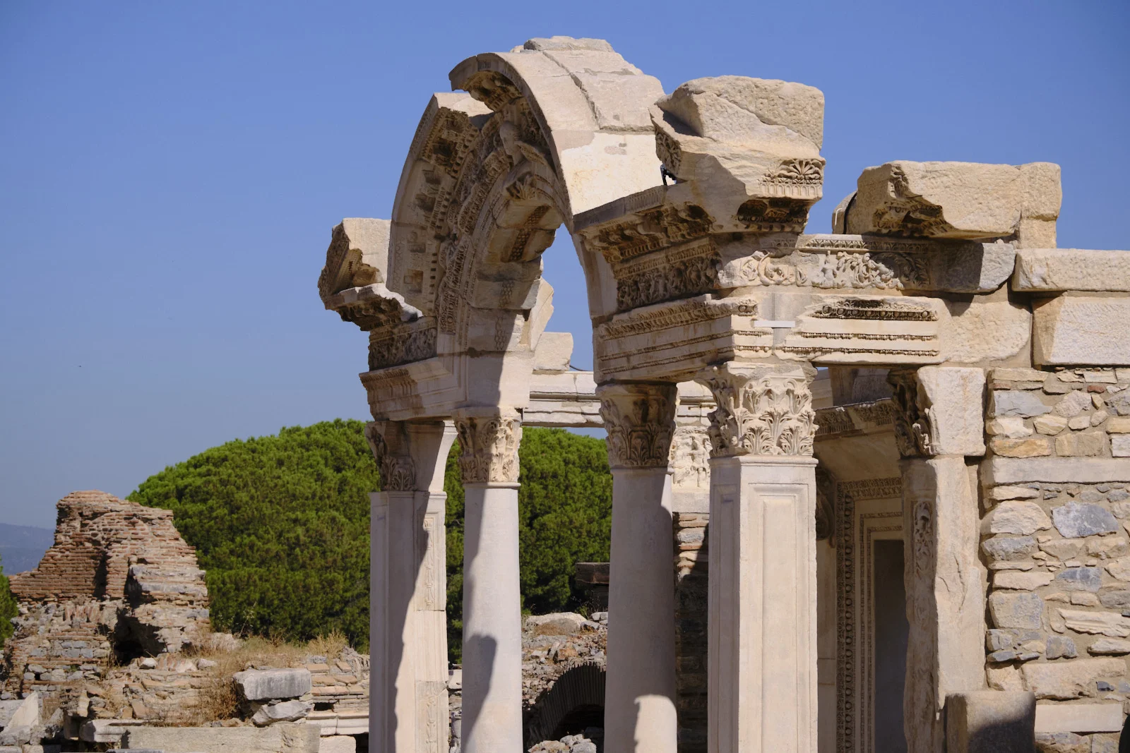 A carved marble arch on Corinthian columns rises among ruins, with a green pine and blue sky behind.