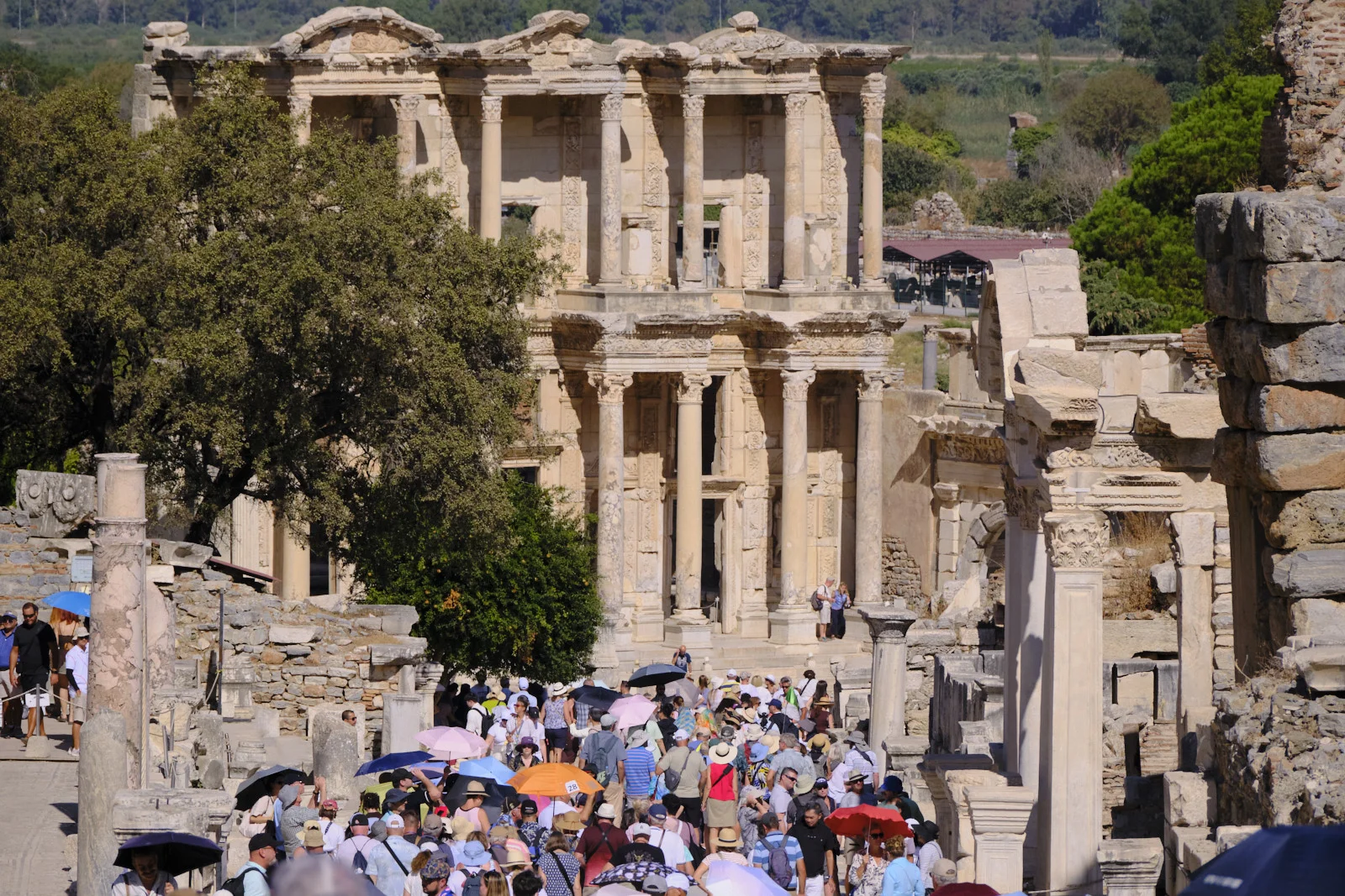 A large crowd with umbrellas fills a ruined street leading to the columned facade of the Library of Celsus.