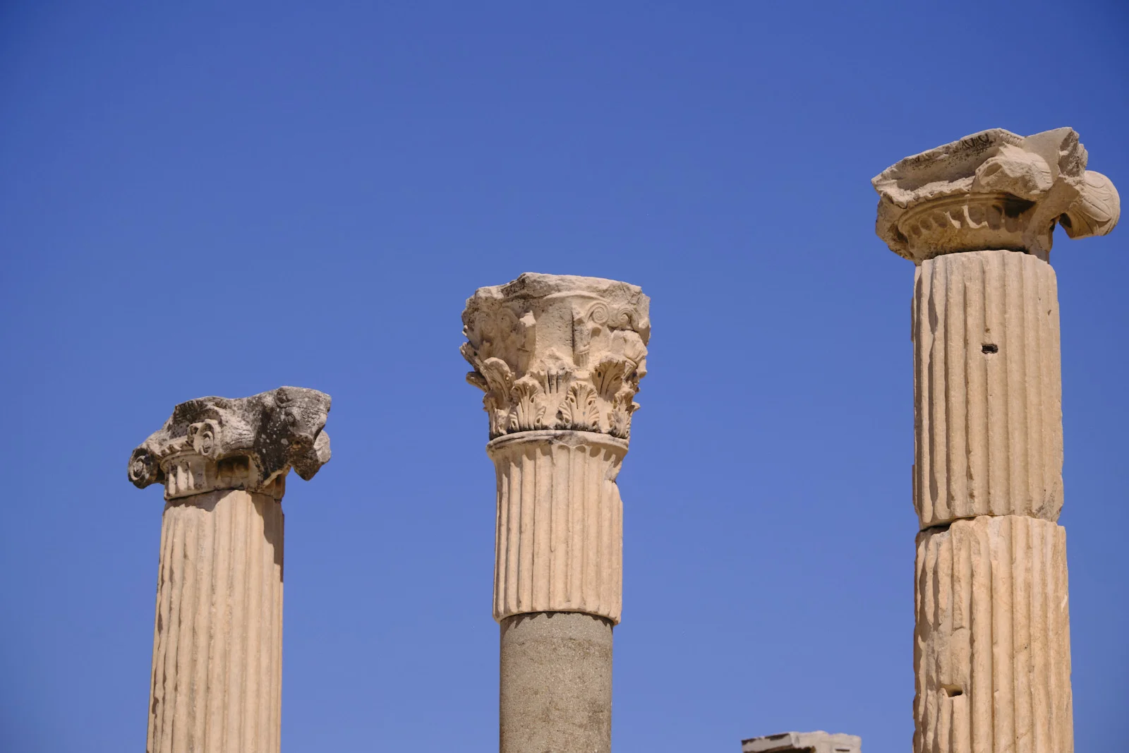 Three ancient fluted columns with damaged Corinthian capitals rise against a clear blue sky.