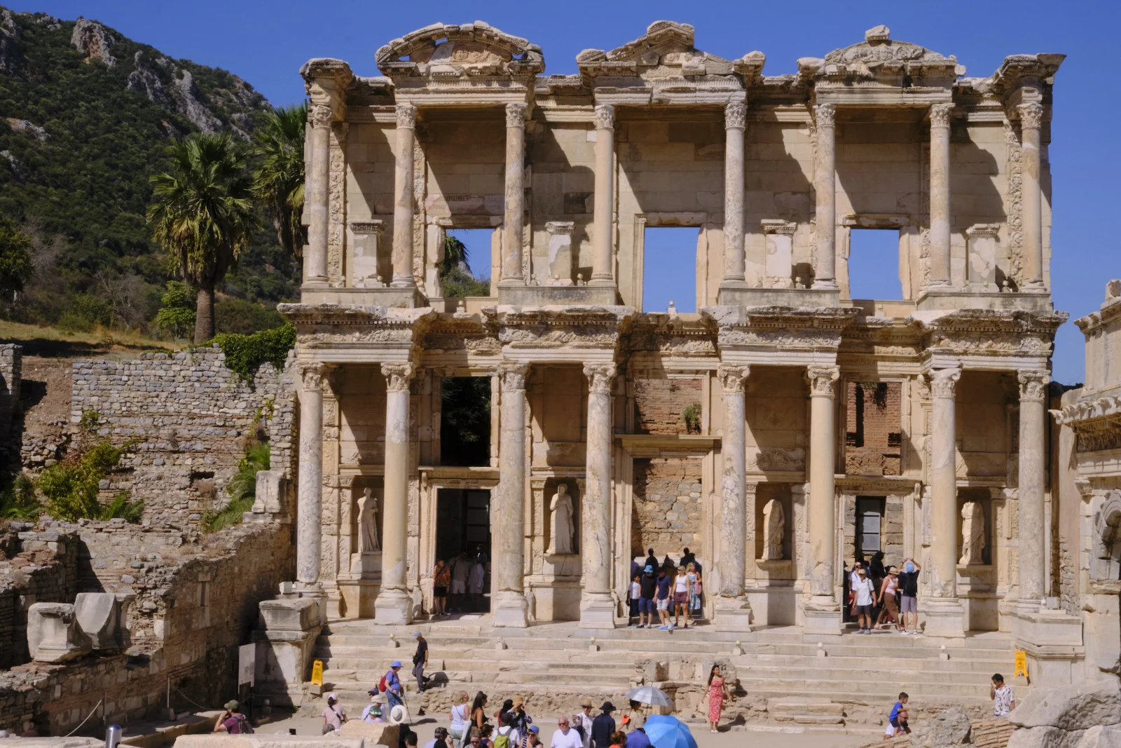 Frontal view of the Library of Celsus facade with statues in niches, columns, and visitors on the wide steps.