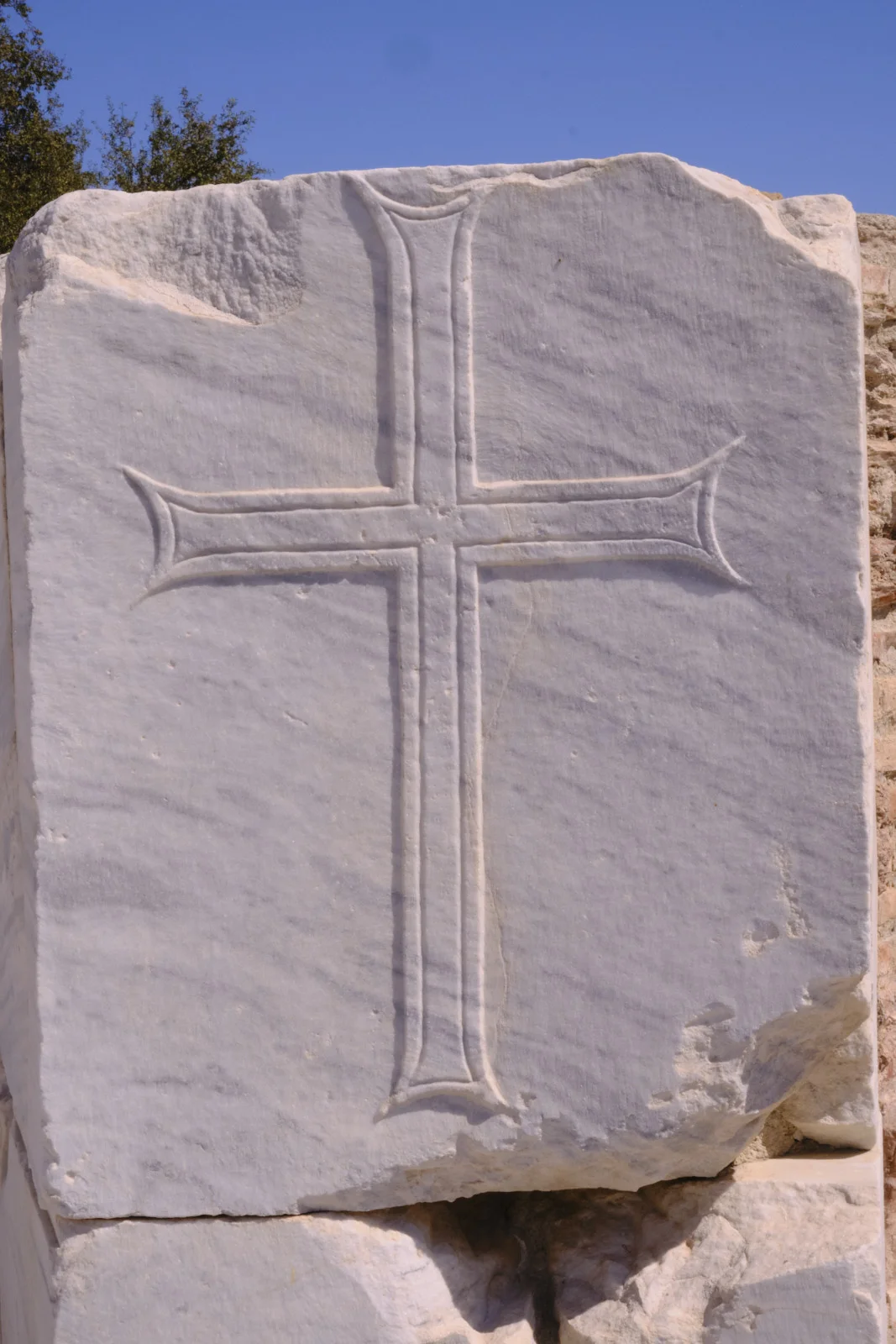 A carved cross with widened ends fills the face of a weathered white marble block against a blue sky.