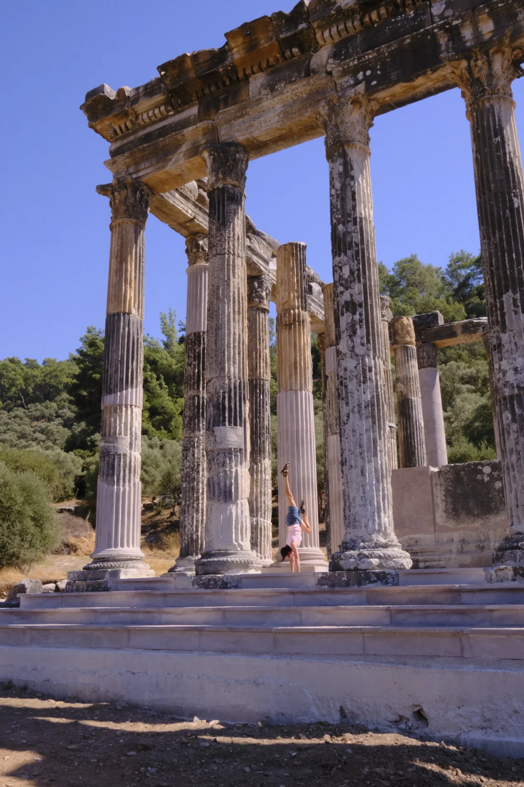 A person in a pink top and denim shorts does a handstand on the steps among the tall columns of a ruined temple.