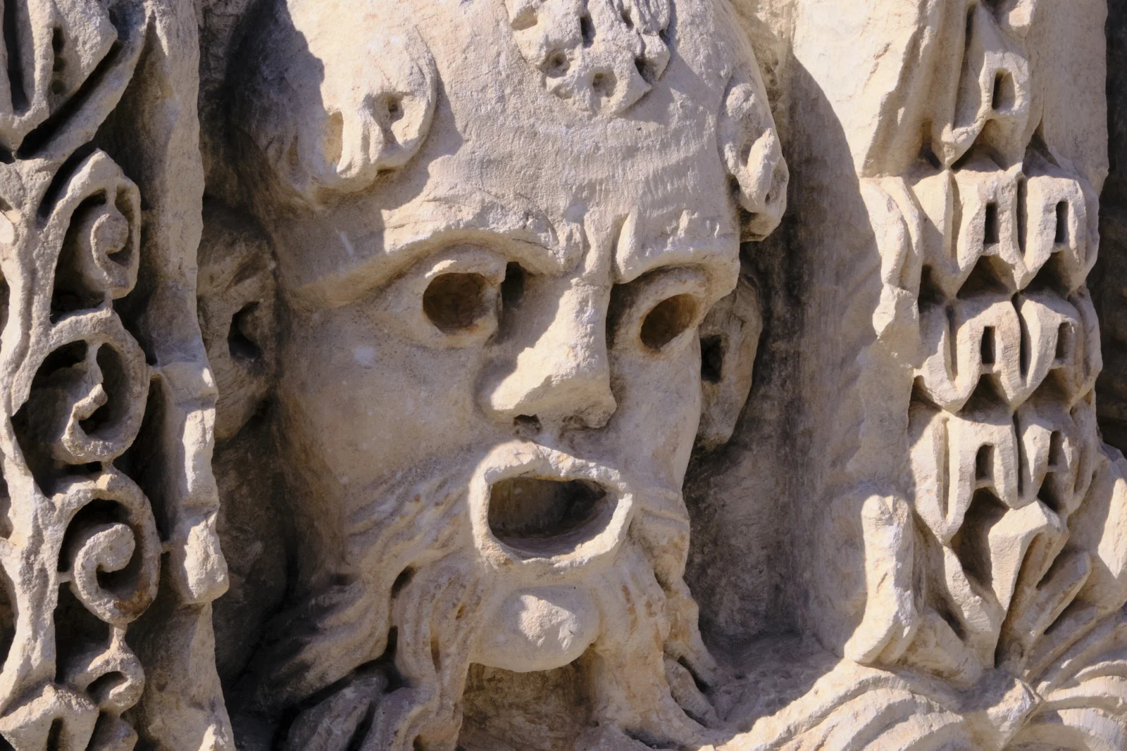 Close-up of a carved limestone mask with hollow eyes, an open mouth, curly hair, and ornamental relief on both sides.