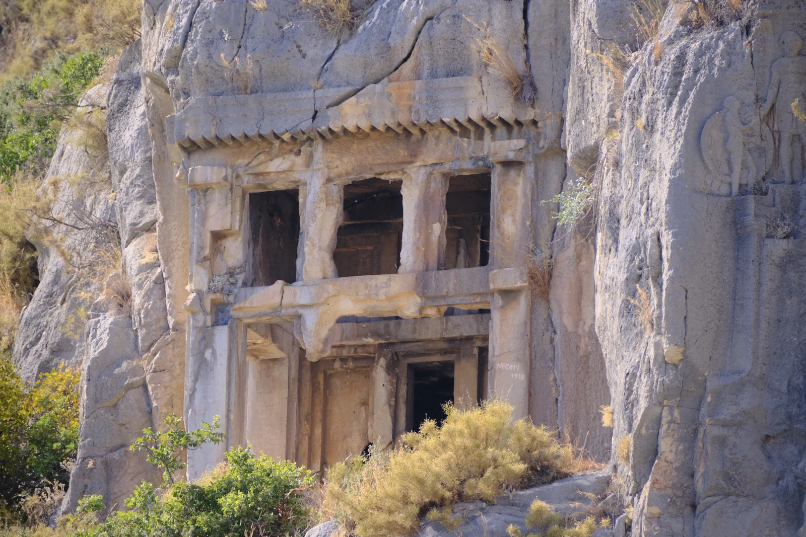 A two-level rock-cut tomb facade with columns and, beside it, a relief of a standing figure holding a round shield.