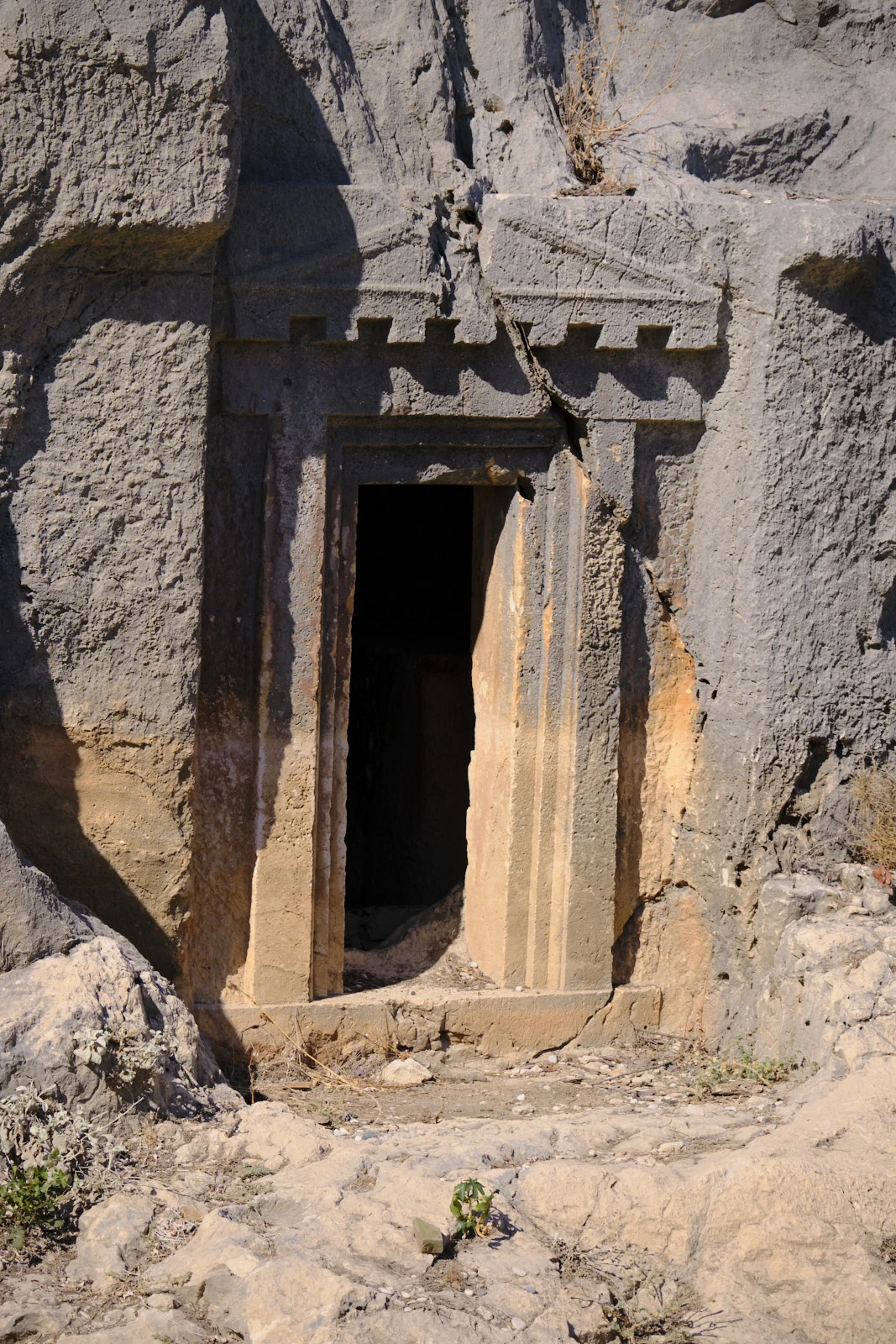 A narrow dark doorway with a triangular pediment carved into a gray rock face above rubble.