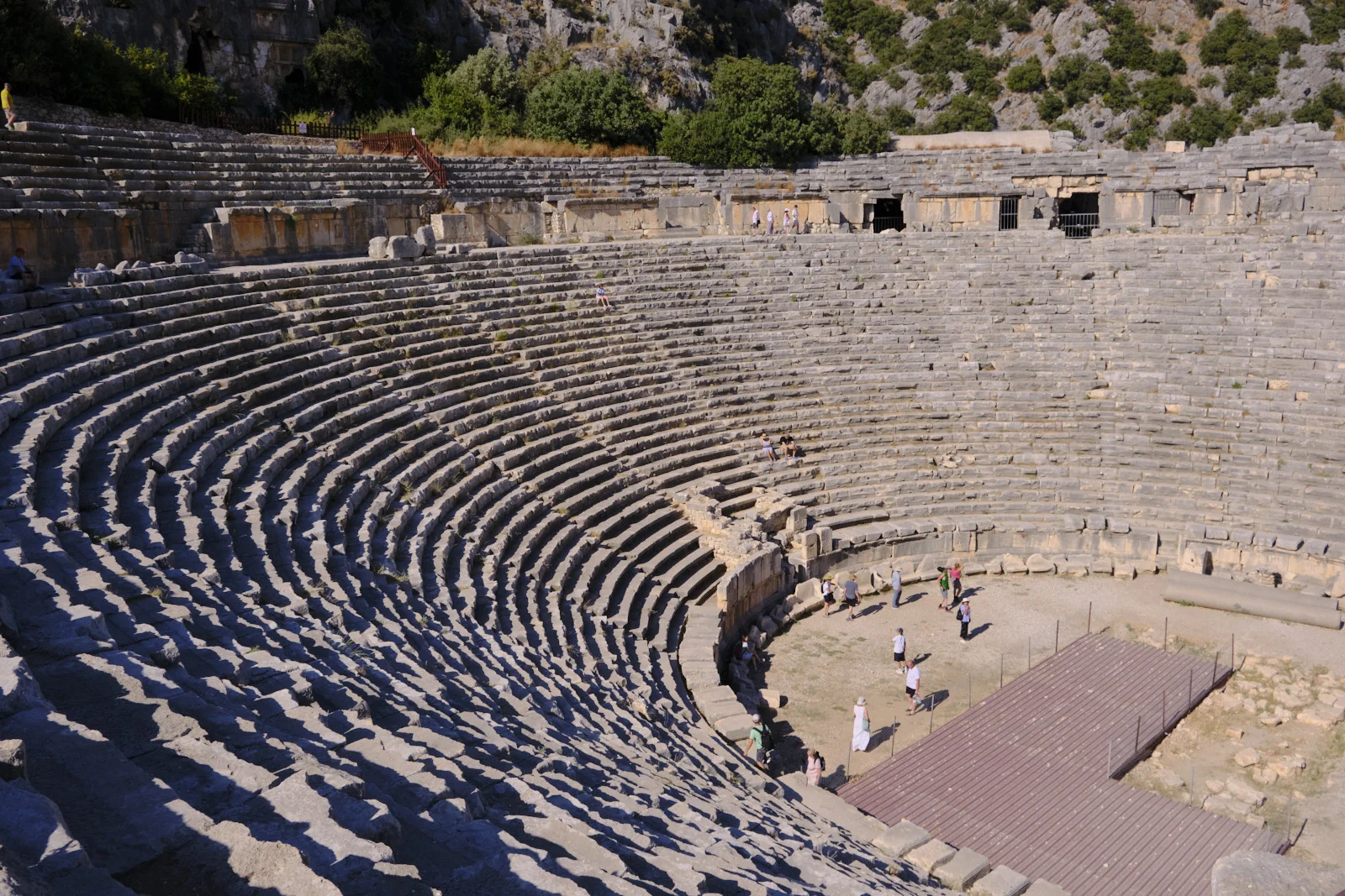 A large ancient theater of curved stone seating with visitors on the floor and a modern wooden platform at the right.