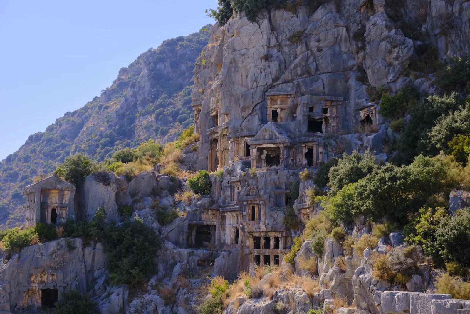 Numerous rock-cut tombs with pediments and dark openings clustered on a cliff, with a green mountain behind.
