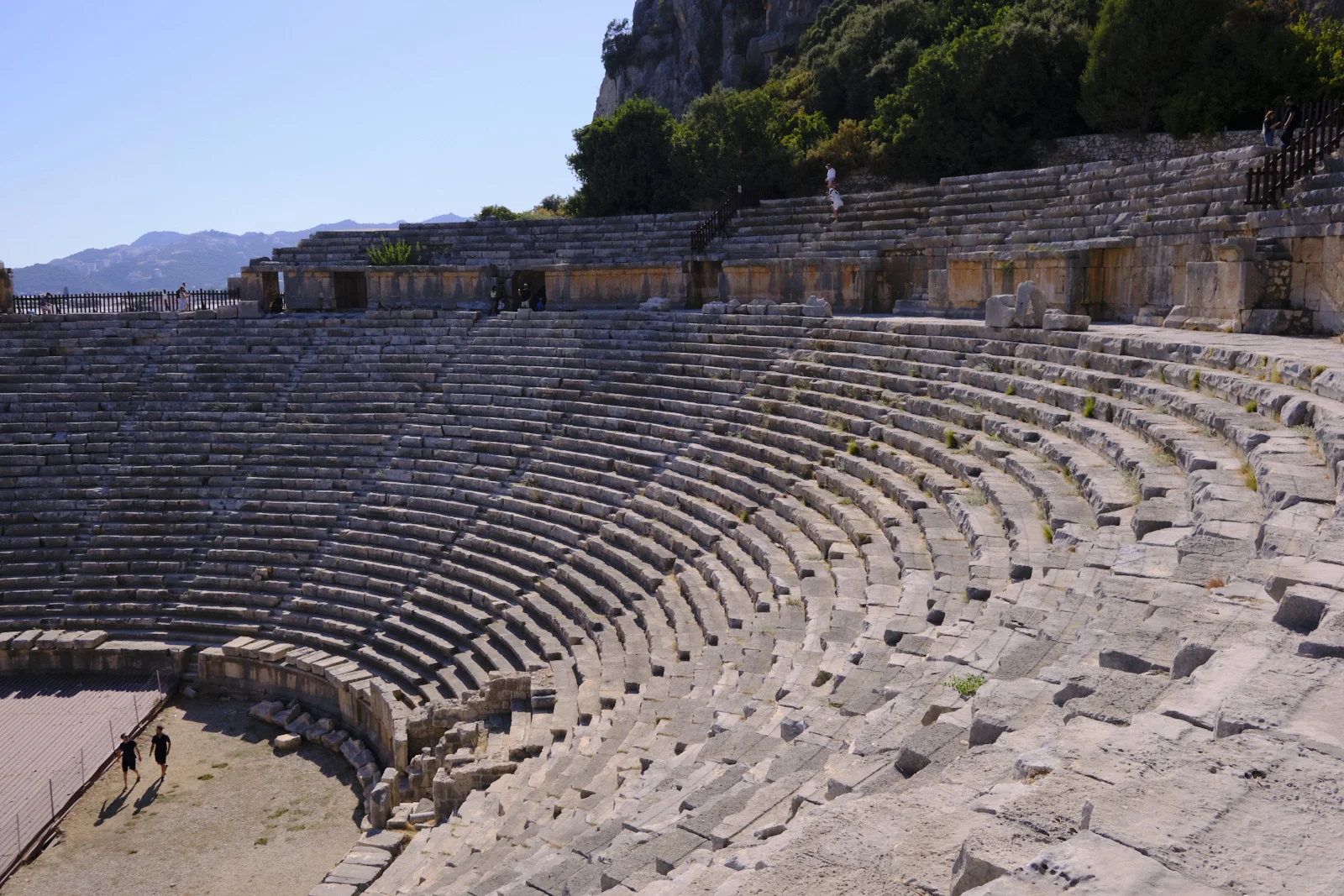 Wide view of an ancient theater's curved stone seats, mostly empty, with two people walking on the open floor.