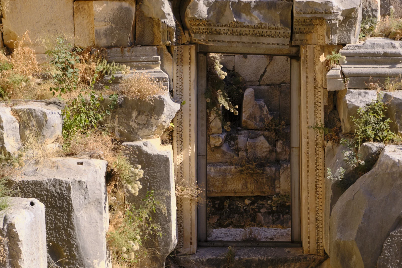 An ornate stone doorway surrounded by collapsed blocks and weeds, with rubble visible through the opening.