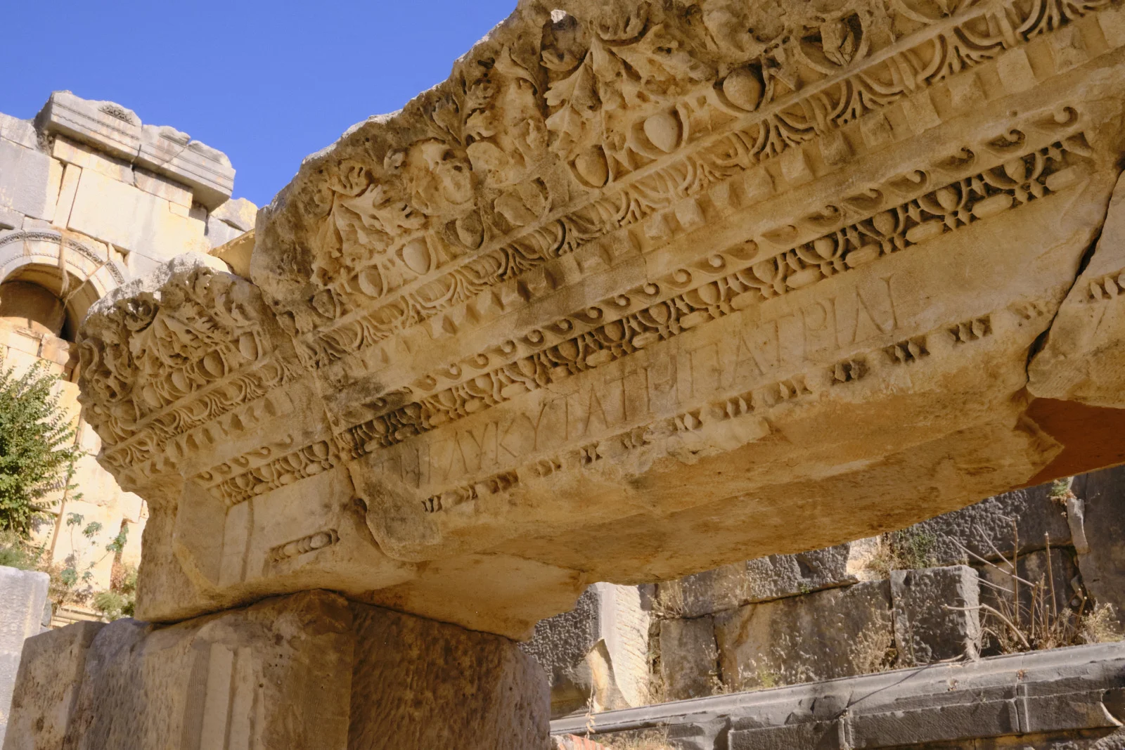 Low-angle view of an elaborately carved stone lintel with an inscription, against a blue sky and ruined masonry.