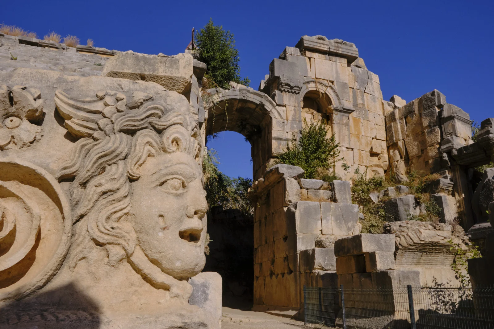 A carved profile of a face with flowing hair beside a stone arch in a ruined theater wall under a deep blue sky.