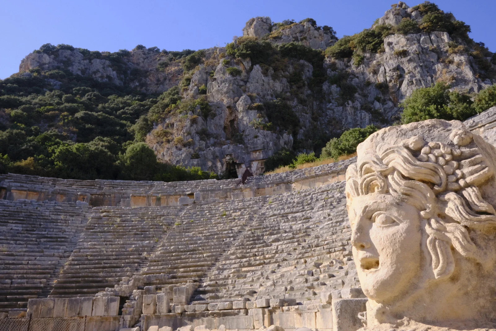 A carved stone head in the right foreground with rows of theater seats and a rocky, tree-covered cliff behind.