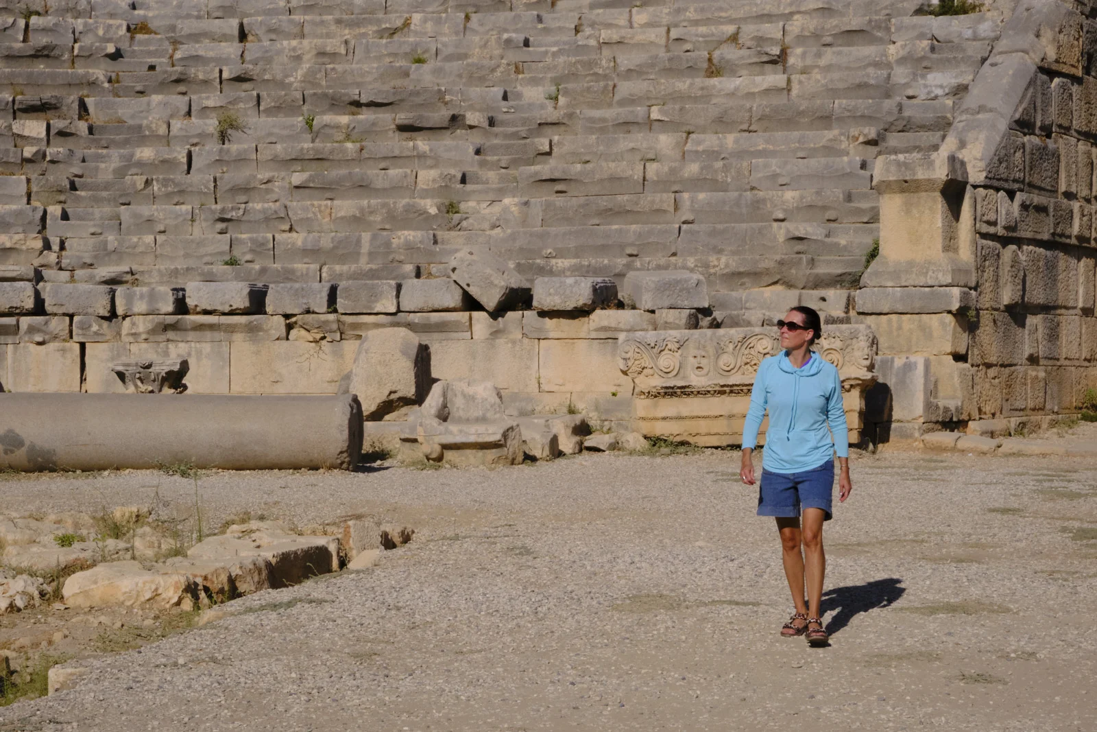 A woman in a blue hoodie, shorts, and sandals walking across gravel in front of ancient theater seating and a fallen column.