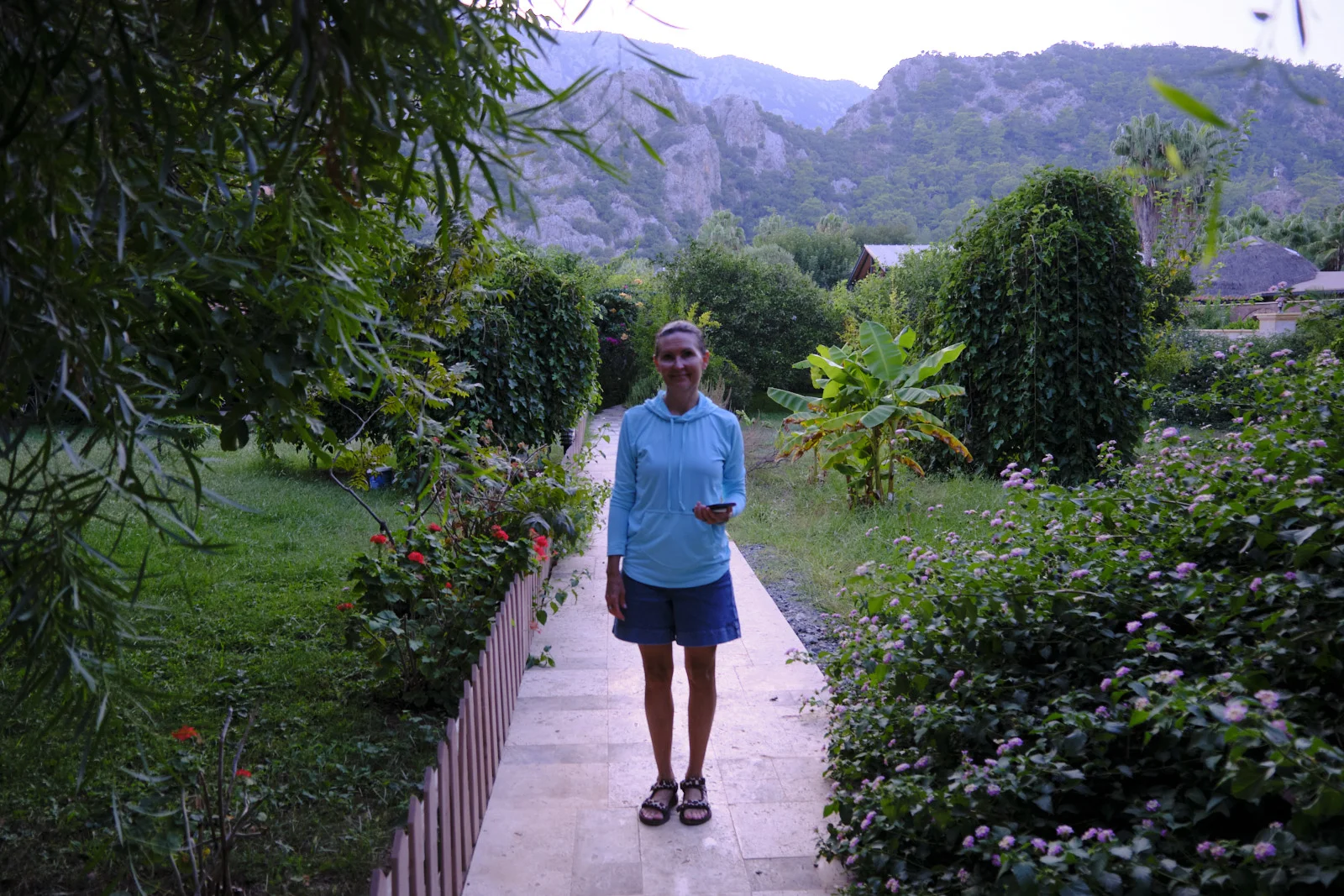 A woman in a blue hoodie and shorts standing on a stone garden path lined with flowers and hedges, mountains behind her.