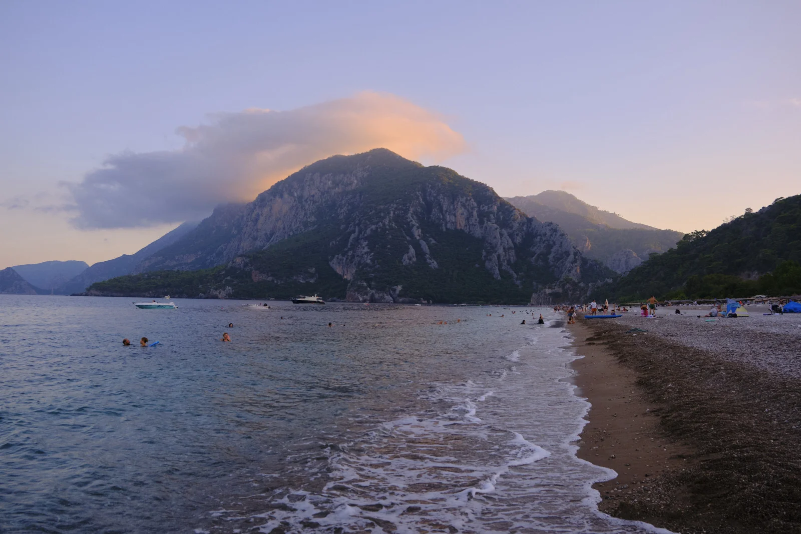 A pebble beach at dusk with small waves, people swimming, boats on the water, and a large mountain under a pink-lit cloud.