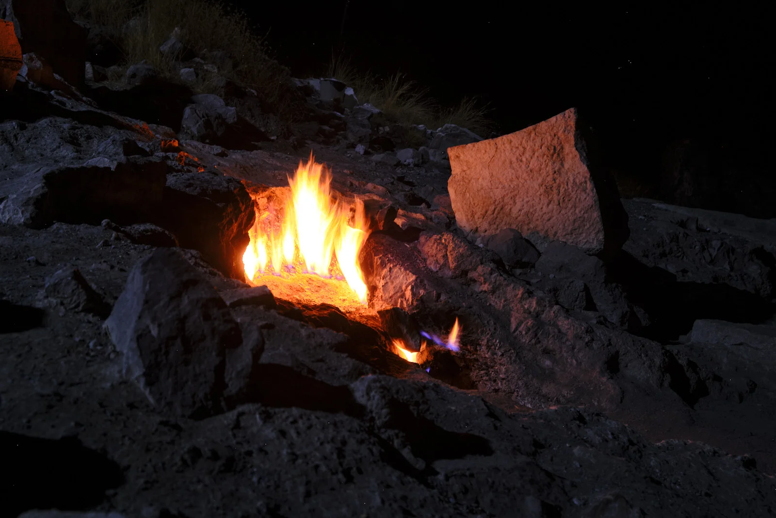 Night photograph of a yellow flame rising from a rocky crevice, with a smaller blue flame and a large stone lit by the fire.