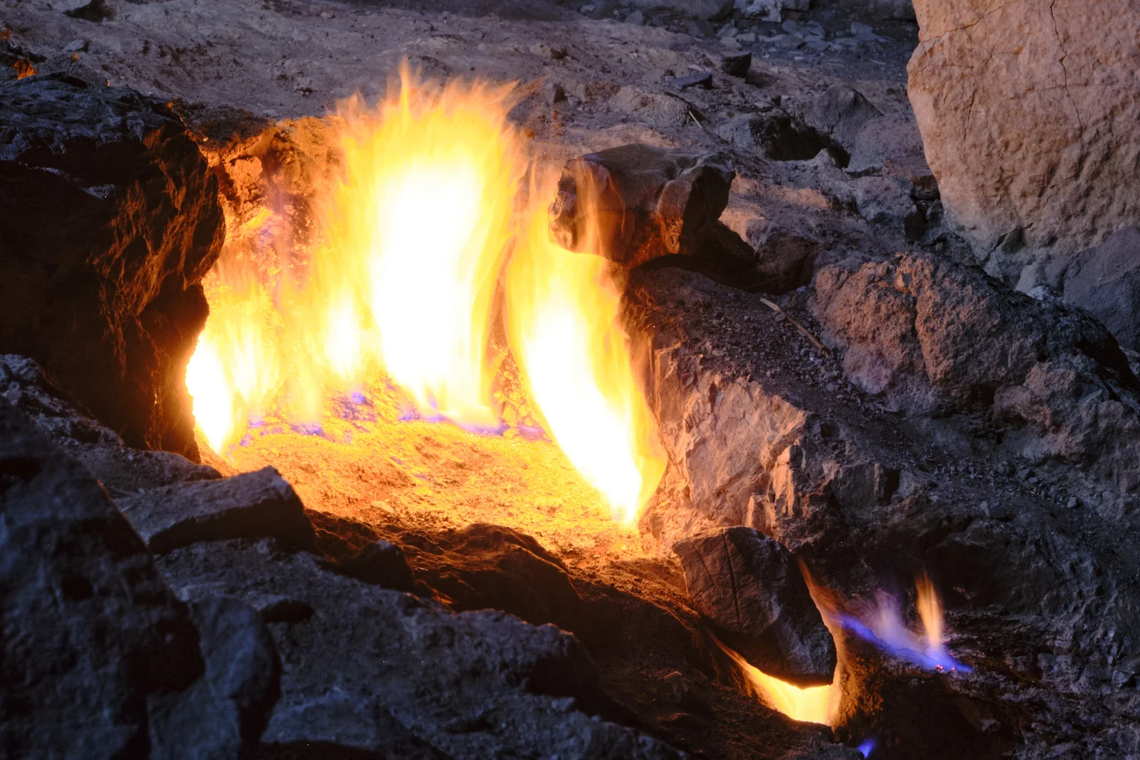 Close view of bright yellow flames erupting from a hollow in dark rock, with blue at the base and a smaller flame below.