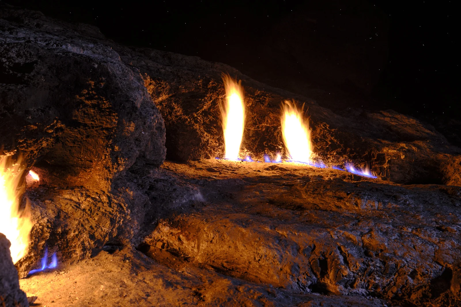 Several tall flames and smaller blue jets burning from a rocky ledge at night, with stars visible overhead.