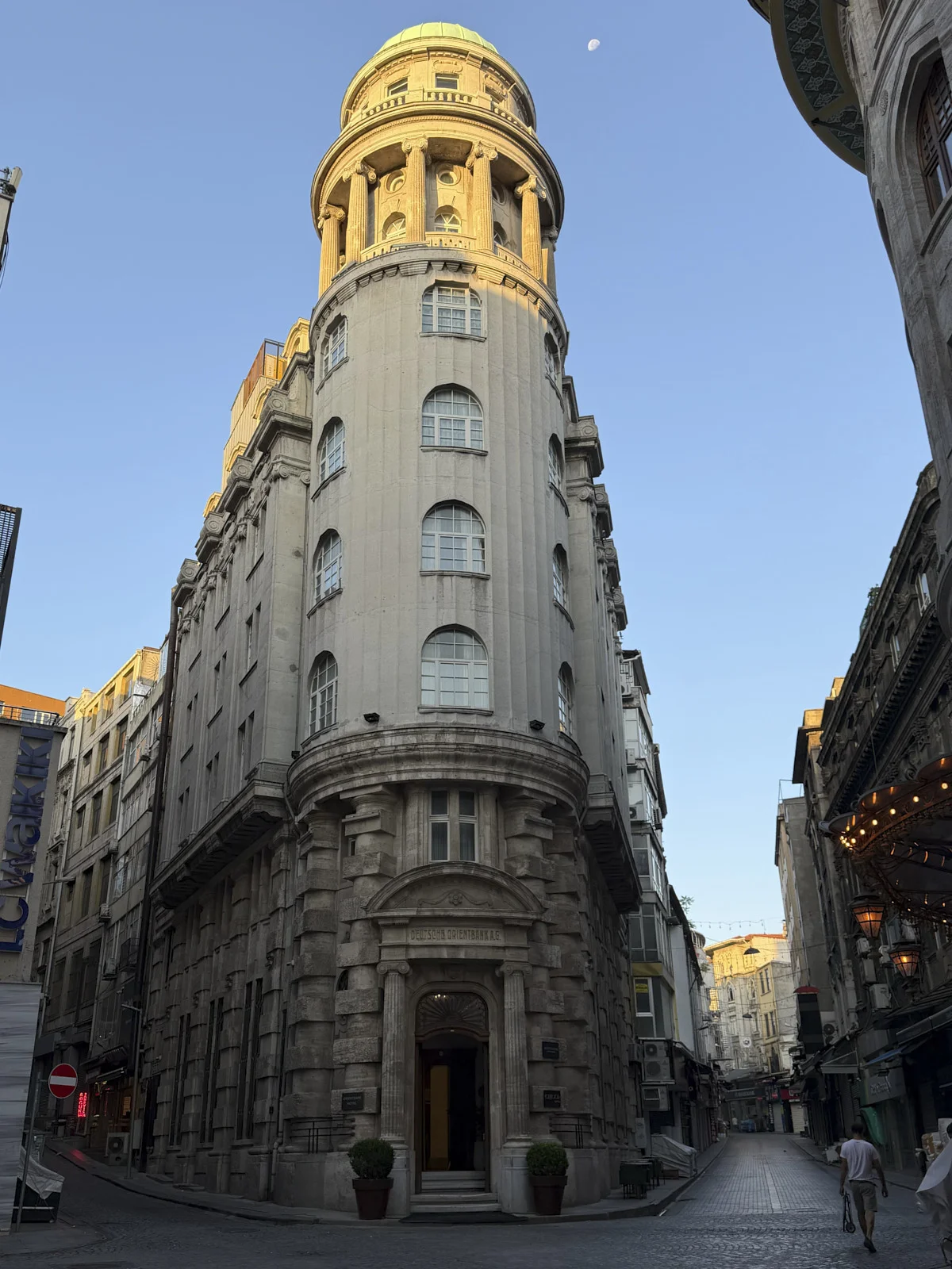 A curved stone building with a gold-lit upper drum and a crescent moon in a clear blue sky, on a cobbled Istanbul street.